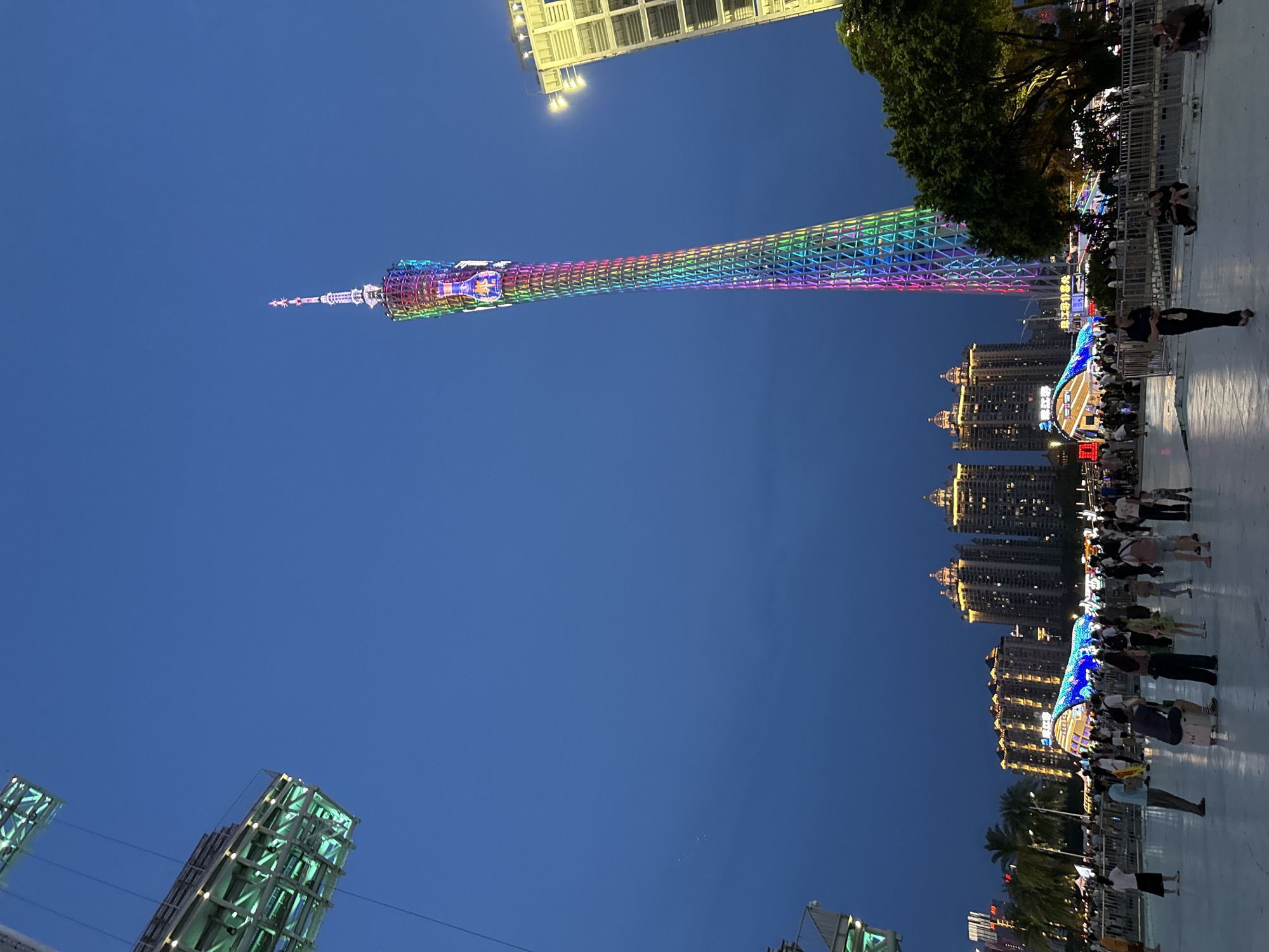 The image captures a nighttime scene in Tianhe District, China, featuring the Canton Tower illuminated with a vibrant rainbow spectrum of lights. The tower dominates the skyline, its intricate lattice structure clearly visible against the deep blue evening sky. To the right of the tower, a tall building with bright yellow lights stands prominently. In the foreground, a wide, paved area is populated by a diverse group of people, many of whom are casually strolling or gathered in small groups. Some individuals are seated on the ground. Behind the crowd, there are illuminated architectural structures and palm trees, suggesting a public gathering space or entertainment district. Above and to the left, a portion of a futuristic, illuminated structure, possibly a suspended walkway or observation deck, is visible, also lit with green lights. The overall atmosphere is lively, indicative of a popular urban attraction at dusk or night.