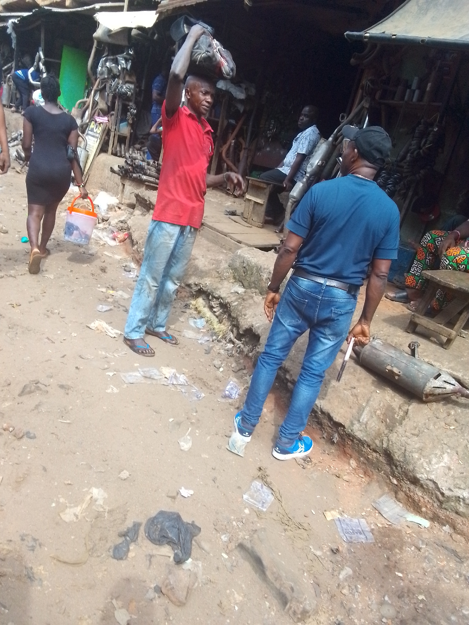 This daytime image depicts a busy street or market scene in Onitsha, Nigeria, characterized by an unpaved, dusty, and debris-strewn ground. The environment suggests an auto parts or repair market due to the numerous metal components visible.
In the foreground, two men and a woman are prominent. On the right, a man wearing a blue t-shirt, blue jeans, a black baseball cap, and blue sneakers stands with his back to the camera, holding a metallic pipe or rod in his right hand. To his left, another man in a red polo shirt and light blue, possibly paint-stained, jeans walks towards the left, balancing a dark bag or bundle on his head. He wears flip-flops. Further left, a woman in a black dress walks away from the camera, carrying an orange plastic bucket, which appears to contain goods like fish, in her right hand. She wears brown sandals.
The background is filled with makeshift market stalls or workshops, constructed from simple frames and roofing materials. These stalls are packed with various metallic objects, strongly resembling vehicle exhaust systems, mufflers, and other auto components. Within these stalls, a man in a light blue patterned shirt is seated on a wooden stool on the right side, looking to the left. Another person is partially visible sitting further back.
The ground is uneven and littered with plastic wrappers and other debris. The overall atmosphere is one of functional commerce and daily activity. Visible text is minimal and largely unreadable due to focus and size, with only faint letters on wrappers and a partial "POS" sign discernible in the background.