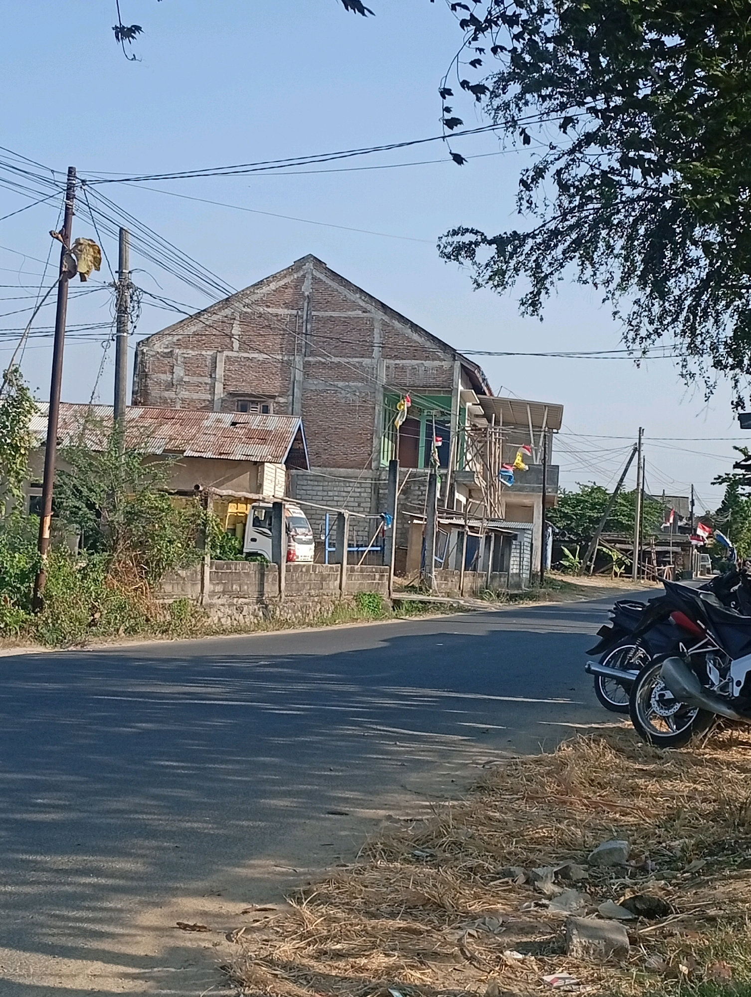 This is a street-level photograph taken in the city of Bonjor, Indonesia. The scene depicts a road with a paved surface and a dry, grassy shoulder. On the left side of the road, there is a low concrete wall bordering a property with dense vegetation. Behind the wall, a small white van is partially visible, parked next to a building with a corrugated metal roof. Further back, a multi-story brick building is under construction or partially renovated, with exposed brickwork and scaffolding. Several electrical poles with a dense network of wires are prominent in the background. The right side of the road shows two parked motorcycles, one black and red, the other black and white. The sky is clear and blue, and sunlight casts shadows on the road. The overall impression is of a semi-urban or rural setting with ongoing construction and everyday street activity.