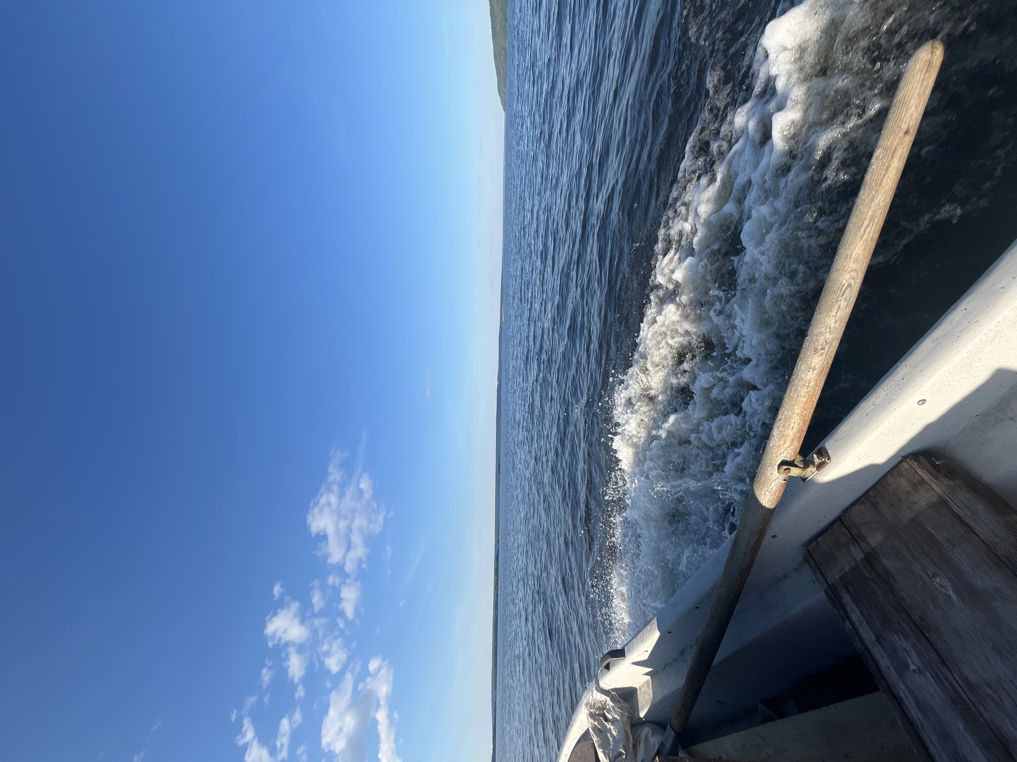 This is a tilted, eye-level shot from a boat moving on the water, with the horizon line running vertically. The scene is split between a bright blue sky and the dark, rippling water. A white boat is partially visible on the right, with a wooden oar or pole extending diagonally across the frame. The water churns with white foam and spray, indicating movement. Scattered white clouds dot the sky. The overall impression is of a sunny day, likely midday, with calm but moving water. The image conveys a sense of travel and the open air. No people are visible. The location is Siukterka, Russia.