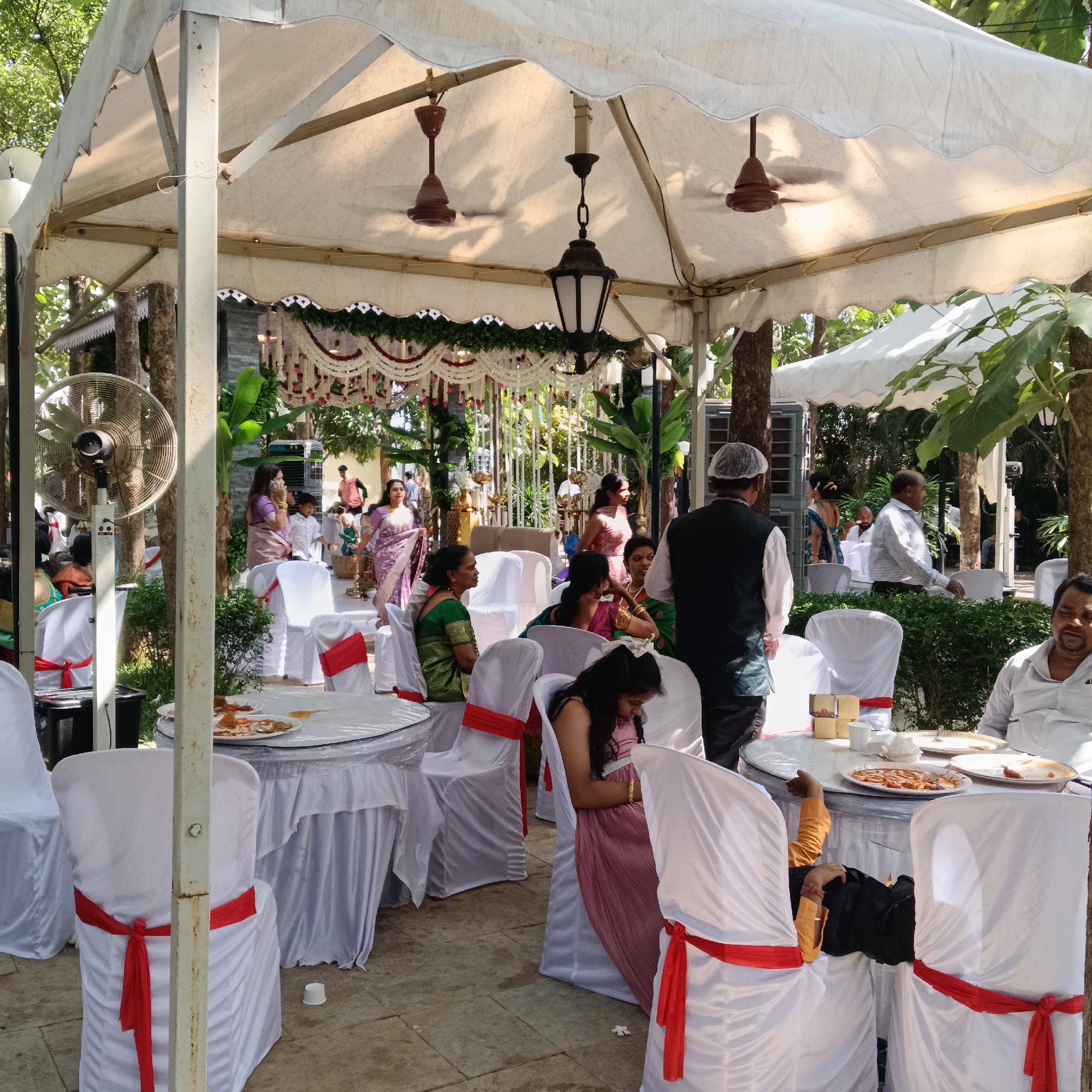 The image depicts an outdoor event, likely a wedding reception, under a white tent structure in Mapusa, India. Several round tables draped in white tablecloths are surrounded by white chairs decorated with red sashes. People are seated at the tables, some eating and talking, while others are standing and interacting. One man is standing wearing a white chef's hat and a black vest. Decorations, including floral arrangements and garlands, adorn the background. Additionally, there are a vintage lantern hanging from the tent's ceiling and several fans that keep the space cooler.
