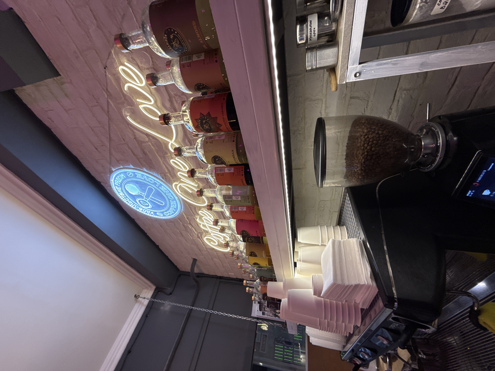 This image depicts an interior shot of a coffee shop, likely in Moscow, Russia, given the presence of Russian text on a label. The foreground shows a black espresso machine with a stack of white paper cups and napkins on top. To the right, a clear glass coffee bean grinder is filled with beans. A shelf above displays an array of colorful bottles, presumably syrups or liqueurs, lined up against a white brick wall. A prominent neon sign illuminates the wall with the words "Coffee One Love" in a glowing script. Further to the right on the shelf are metal containers, one of which has a label with Russian text that appears to read "Мускатный орех" (Nutmeg). The overall ambiance is warm and inviting, with soft lighting and a focus on coffee-related items.