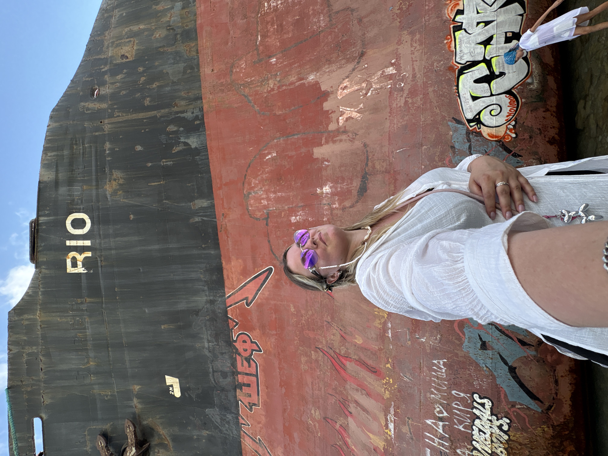 In the city of Kabardinka, Russia, a woman stands in front of a grounded, rusty ship. She wears a white blouse and purple sunglasses. The ship is covered in graffiti, including the word "RIO" in large white letters on the upper section, and various tags and drawings on the red hull. To the right of the woman, a small child in a white outfit walks near the water's edge, also in front of the graffiti-covered hull. The sky above is blue with scattered clouds.