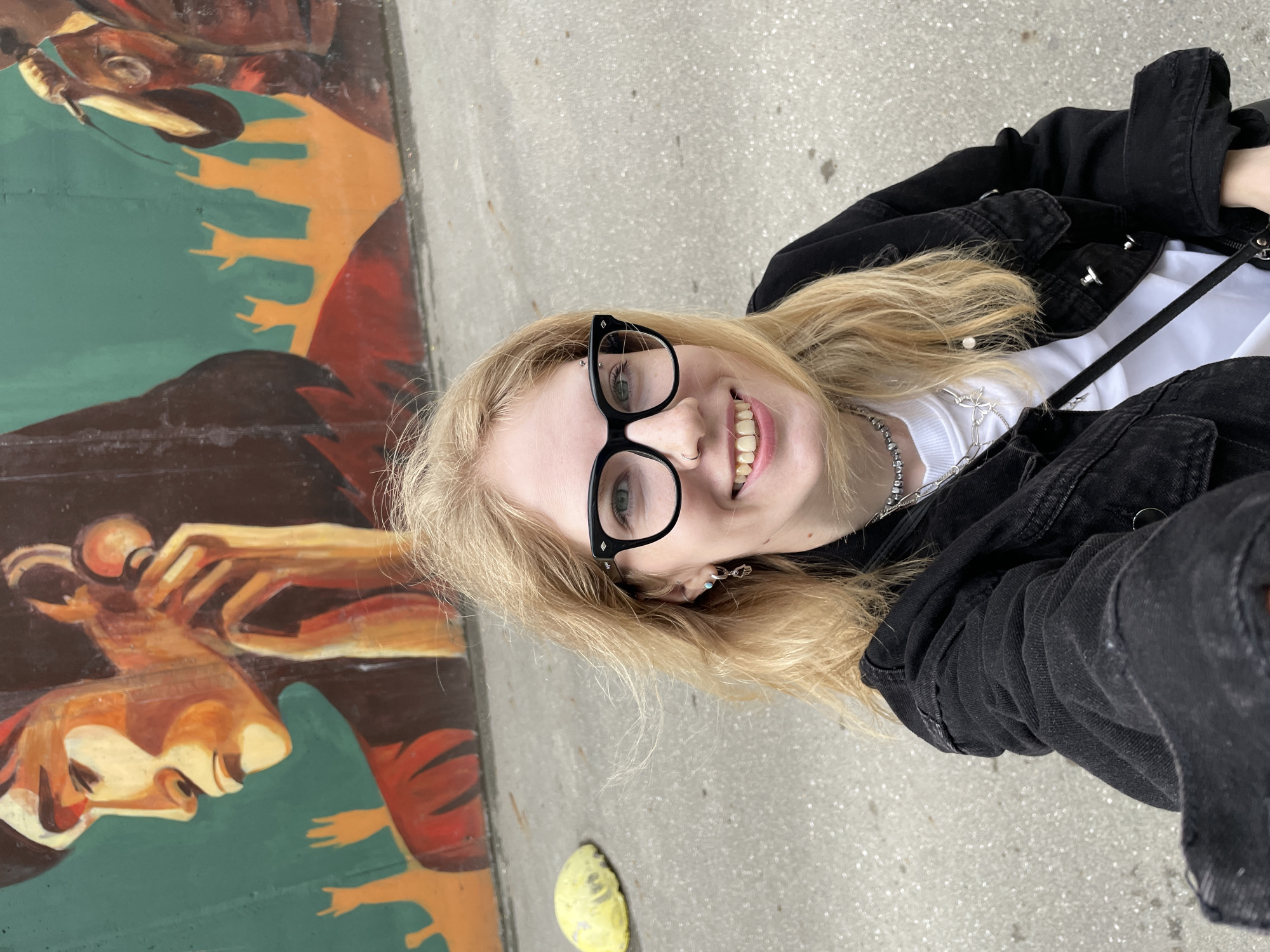 The image is a medium shot selfie of a young woman with blonde hair and glasses, taken outdoors in front of a mural. The mural depicts stylized figures with a predominantly green and orange color palette. The woman is wearing a black denim jacket over a white t-shirt and multiple necklaces. She has a ring in her nose and a piercing above her lip. Her left ear has two piercings, one with a turquoise stud and the other with a silver dangling earring. The background shows a gray, paved ground with a yellow, rounded object on the left side. The lighting is natural, suggesting it is daytime. The location of Izhevsk, Russia is not discernible from the image itself; this information is external.