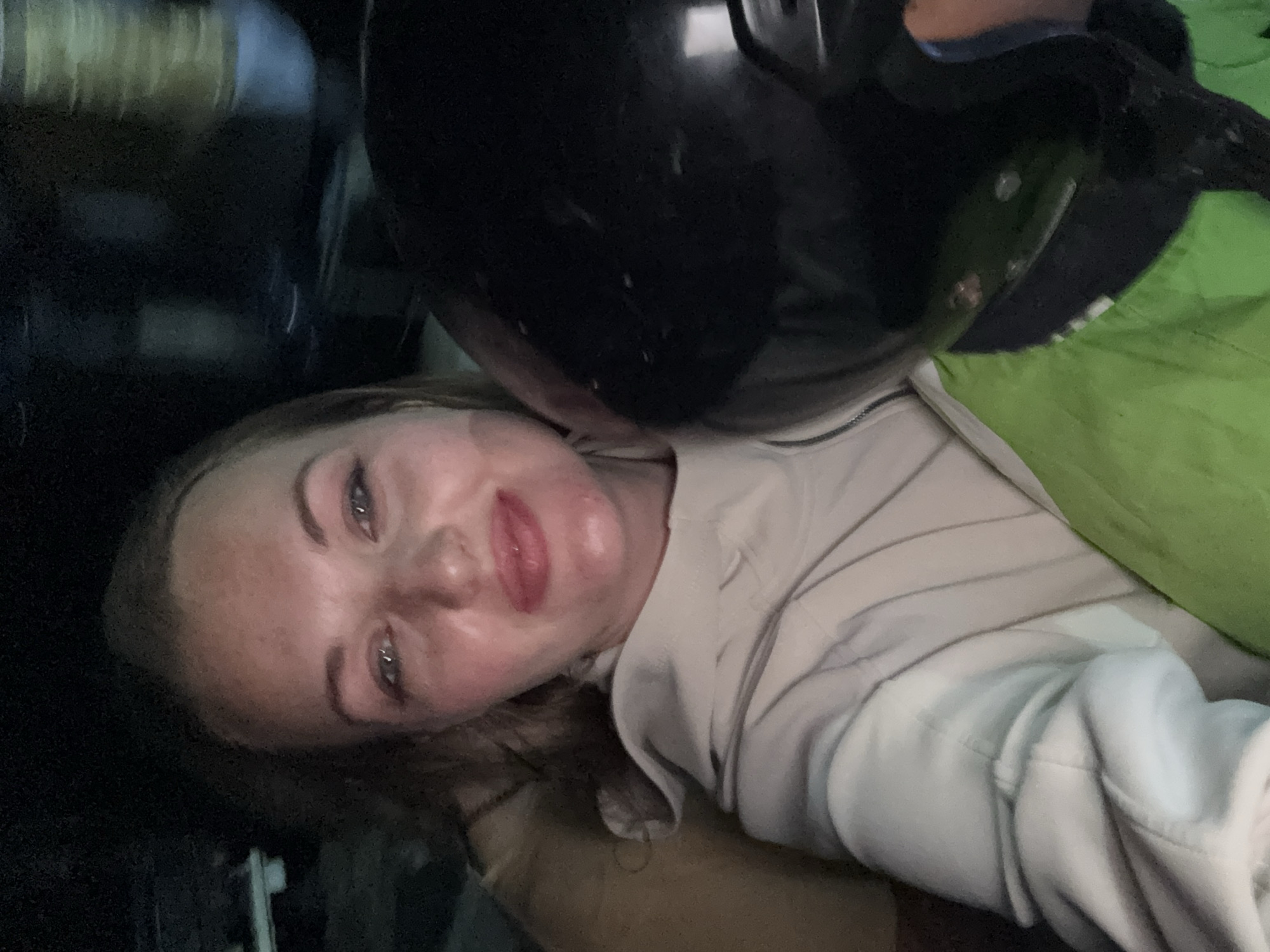 A selfie is taken by a woman at night. She is in a vehicle, likely a scooter, as she is wearing a helmet that is partially visible on the right side of the image. She has blonde hair and is wearing a light-colored hoodie. The background is dark, with some blurred lights suggesting movement. The scene appears to be in an urban environment, possibly Denpasar, Indonesia. The overall lighting is low, contributing to the nighttime atmosphere.