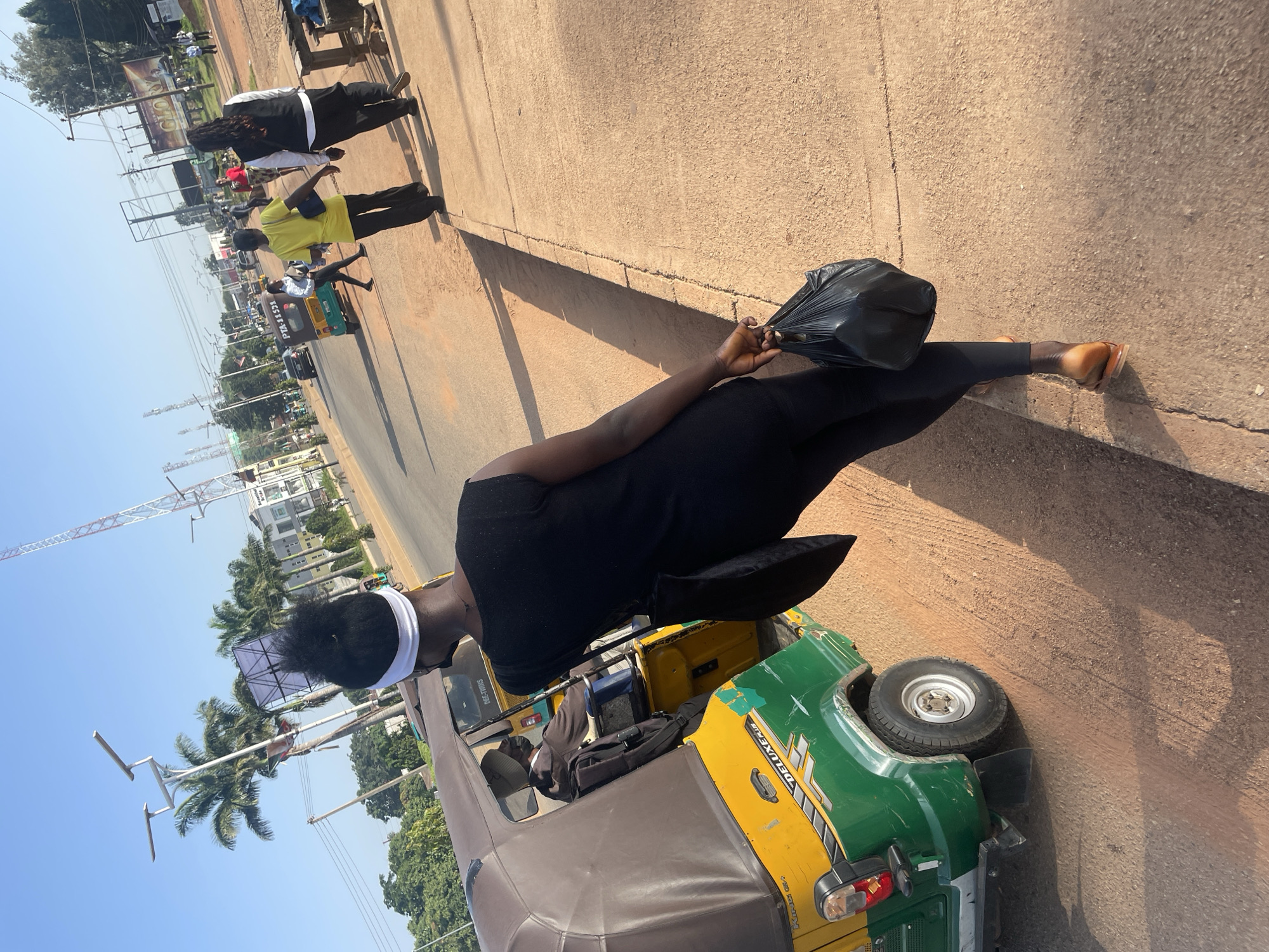 In the city of Jos, Nigeria, an individual walks on a dirt-colored sidewalk adjacent to a road on a clear, sunny day. The person, seen from behind, is wearing a black sleeveless top, black leggings, and brown sandals. They carry a black plastic bag in their right hand. To their left, a green and yellow auto-rickshaw, identified as a "DELUXE PLUS" model, is parked. The rickshaw has a brown canopy and appears to have passengers inside. Further down the road, several other people are visible walking or standing, along with vehicles and buildings. A large billboard with the word "GLORY" is present in the background. The environment suggests a bustling urban or semi-urban setting with elements of local transportation and commerce.