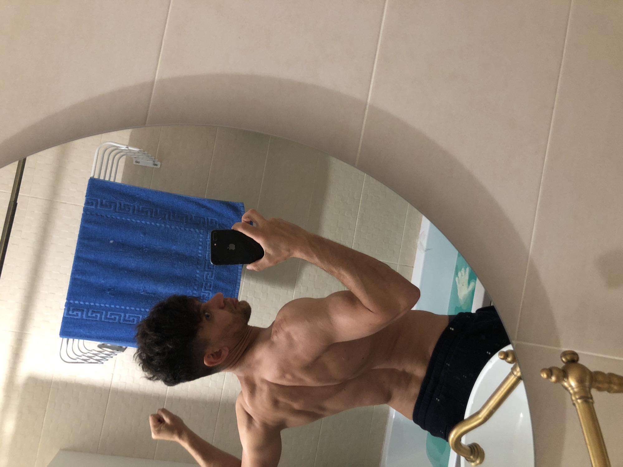 A man is taking a mirror selfie in a bathroom. He is shirtless and wearing dark shorts, flexing his back muscles. A blue towel hangs on a rack behind him, and a bathtub is visible in the reflection. The bathroom appears to be tiled in neutral tones. The image is not specific to any location in the city of Saint Petersburg, Russia.