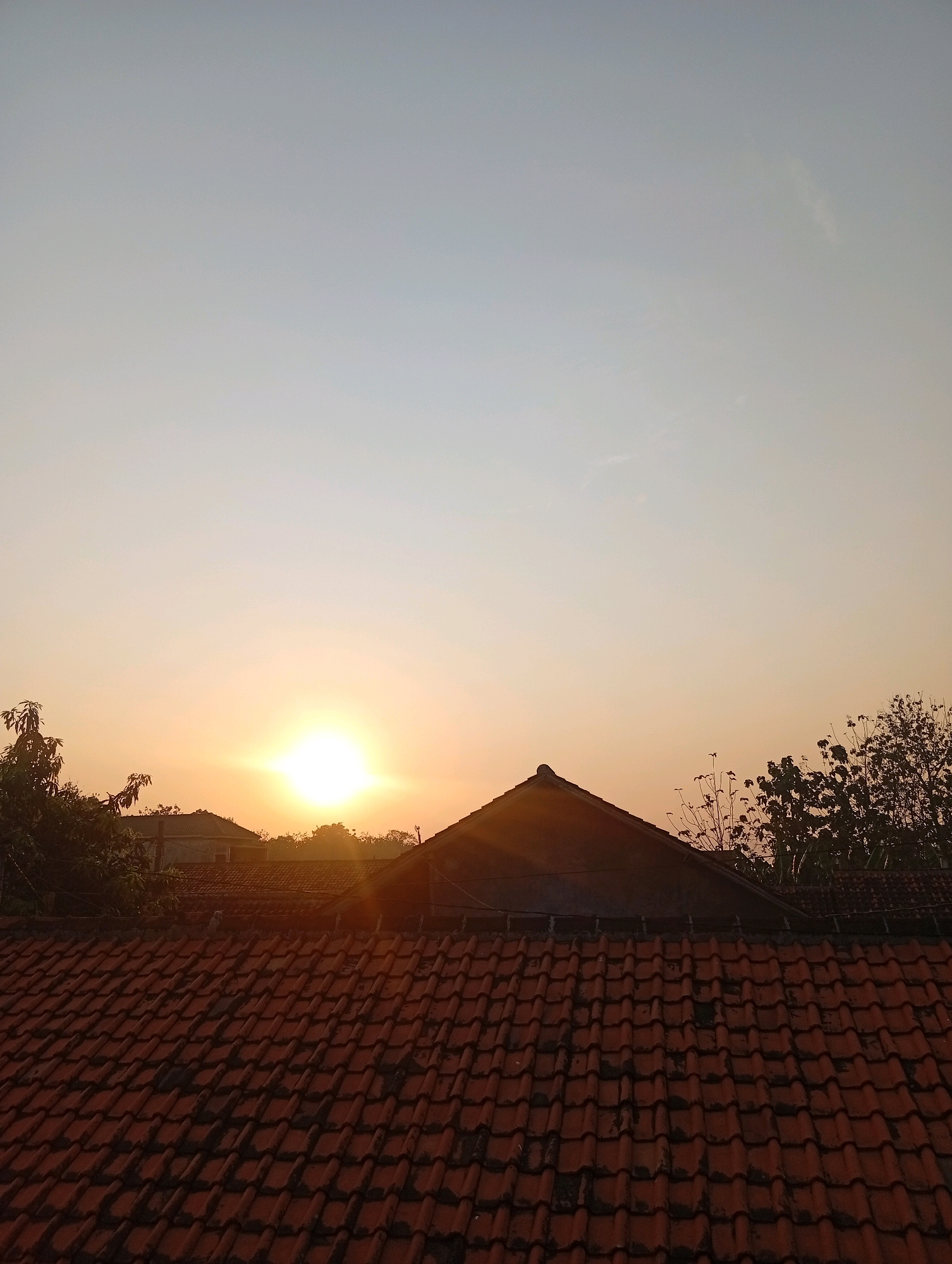 A wide-angle, eye-level shot captures a serene sunrise over a residential area in Kebandingan, Indonesia. The foreground is dominated by a weathered, red-tiled roof, its tiles showing signs of age and grime. Behind the roof, several houses with similar tiled roofs are visible, some partially obscured by trees.
The sky is a gradient of soft colors, transitioning from a pale blue at the top to a warm orange and yellow near the horizon. A bright, radiant sun is the focal point of the sky, its rays casting a warm glow and creating a lens flare effect in the center. Silhouettes of trees and their branches are scattered across the scene, framing the sky and adding depth. The overall impression is one of quiet beauty and the peaceful start of a new day.
