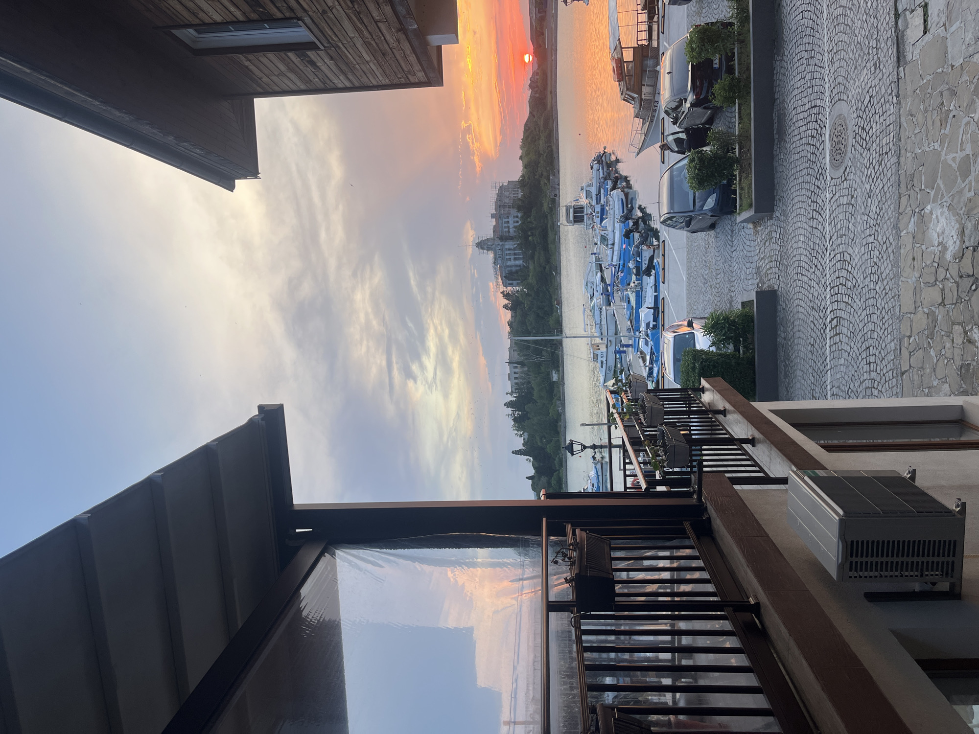 The image captures a picturesque waterside scene at sunset, viewed from what appears to be an elevated balcony or terrace. The dominant colors are the warm oranges and reds of the setting sun, contrasting with the cooler blues and grays of the sky and water.
In the foreground, to the left and bottom, parts of a building are visible, featuring a wooden structure with a window, and a modern balcony with dark metal railings and what seems to be a transparent weather-proof screen. An outdoor air conditioning unit is mounted on the building wall on the right side.
The midground features a body of water, likely a lake or harbor, where numerous small motorboats are docked in neat rows. Most of these boats are white with blue covers or accents. Visible text on some boats includes "VENTURA", "MERCURY", "225 VERADO", and "EVINRUDE", suggesting various engine brands. The water reflects the vibrant colors of the sunset. On the far shore, distant buildings, some appearing under construction, are nestled among trees.
To the right, a paved area with distinct cobblestones, arranged in a fan pattern, extends away from the viewer. Several parked cars, primarily dark-colored, are visible along this path, with some small green bushes in planters separating them from the water. A circular manhole cover is embedded in the cobblestone path.
The background is dominated by the expansive sky, showcasing a dramatic sunset. The sun, a bright orange disc, is low on the horizon, casting an orange glow across the clouds and the water. Faint silhouettes of birds can be seen against the sky. The overall impression is one of a calm and serene evening at a marina or lakeside resort.