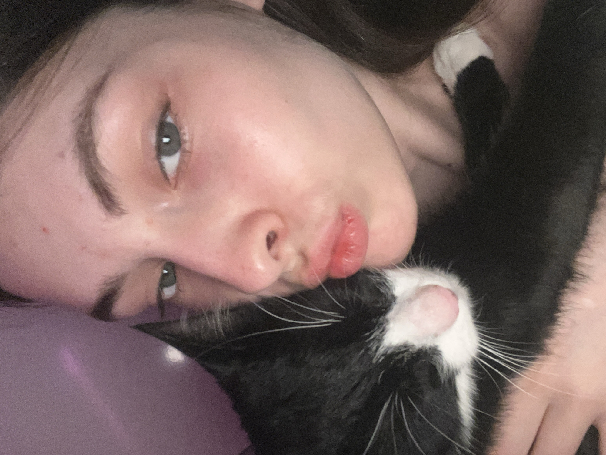 A close-up, slightly angled shot of a young woman lying down, cheek to cheek with a black and white cat. The woman has fair skin, dark hair, and grey eyes. Her lips are slightly pursed. The cat is mostly black with white markings on its face and chest. The background is a soft, lavender color, possibly a pillow or bedding. There is no information given that can be used to determine the location is Saint Petersburg, Russia.