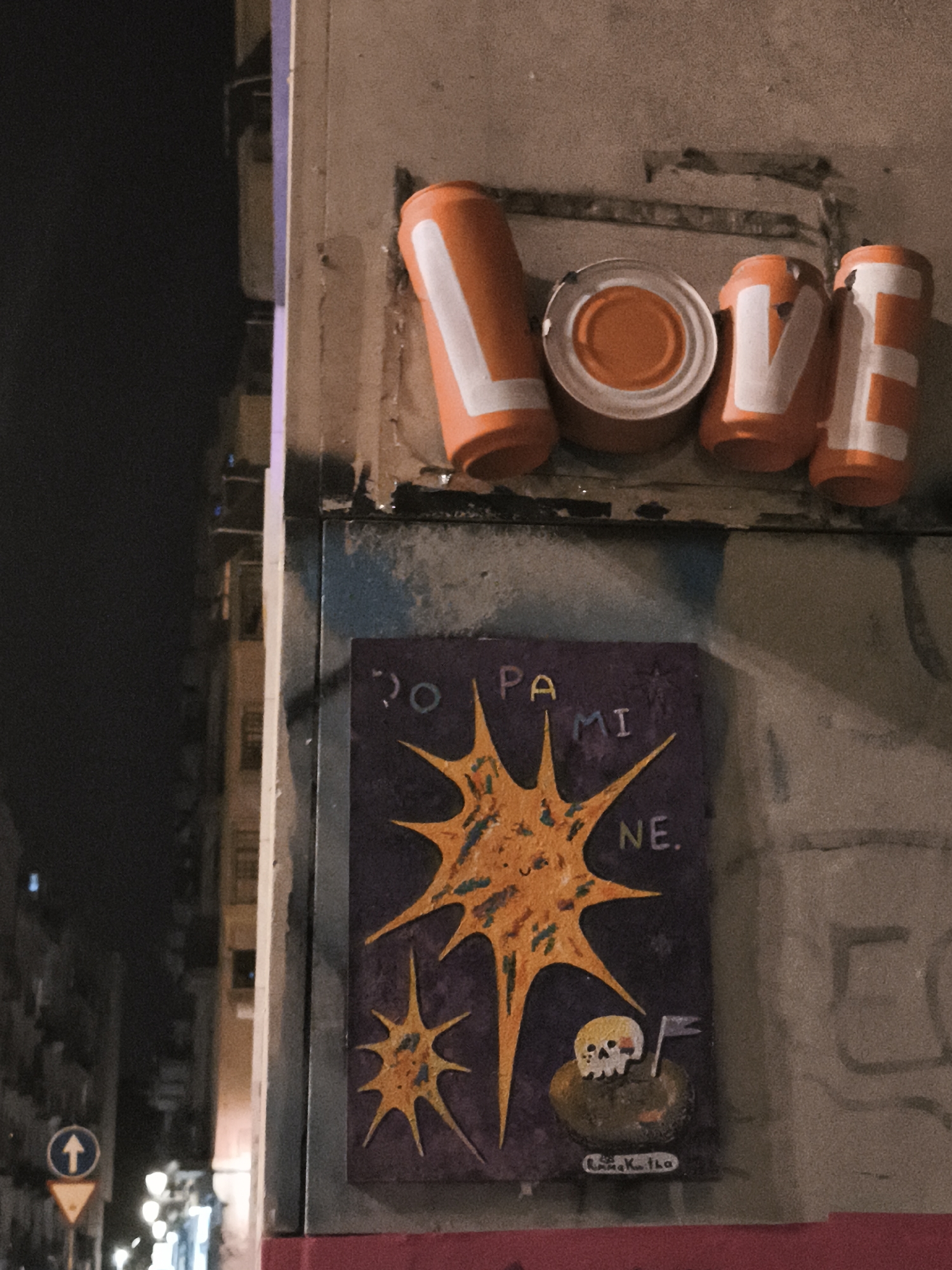 The image captures an urban scene at night. In the foreground, against a concrete wall, are artistic elements. At the top, the word "LOVE" is spelled out with orange-painted tin cans, the letter "O" represented by an intact can. Below this, a purple painted sign reads "DOPAMINE" with a yellow starburst design and a small skull with a flag. In the background, out of focus, is a street with buildings and street lights, likely in Barcelona, Spain. A directional sign is visible on the left.