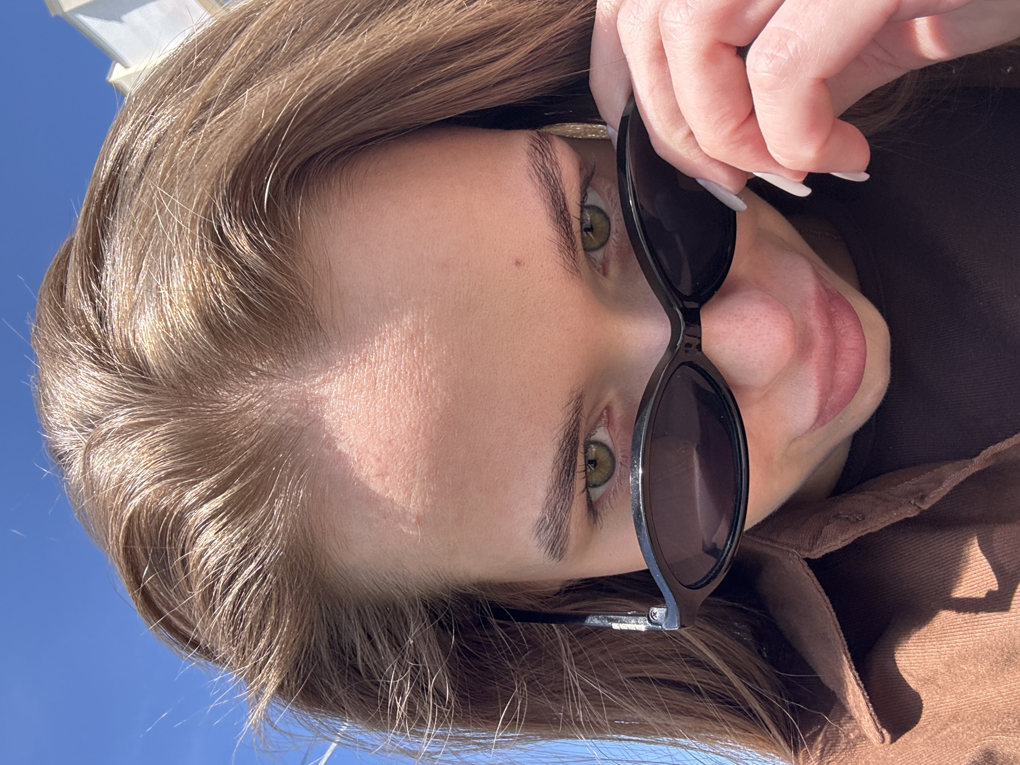 A young woman is holding sunglasses, possibly outdoors on a sunny day, with blue sky visible. Her hair is long and light brown, and she is wearing a brown jacket.
The location is not discernible from the image.
