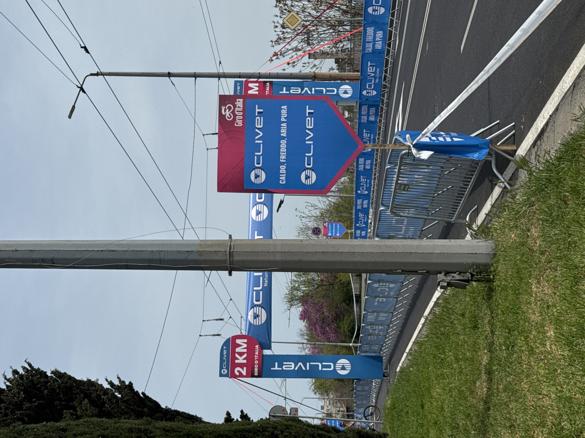 The image shows an outdoor scene, likely along a street during the Giro d'Italia cycling race, with prominent Clivet brand advertisements. A gray concrete utility pole stands in the foreground. Attached to the pole are various banners and signs displaying the Clivet logo and promotional phrases such as "CALDO, FREDDO, ARIA PURA" and indicating "2 KM" to a location. The Giro d'Italia logo is visible on the banners. Along the side of the road, there is a metal barrier, also adorned with Clivet banners. The environment includes green grass and some trees. The location appears to be Burgas, Bulgaria, given the Cyrillic alphabet in the background, and information in the OCR. The sky is overcast, suggesting it might be a cloudy day.
