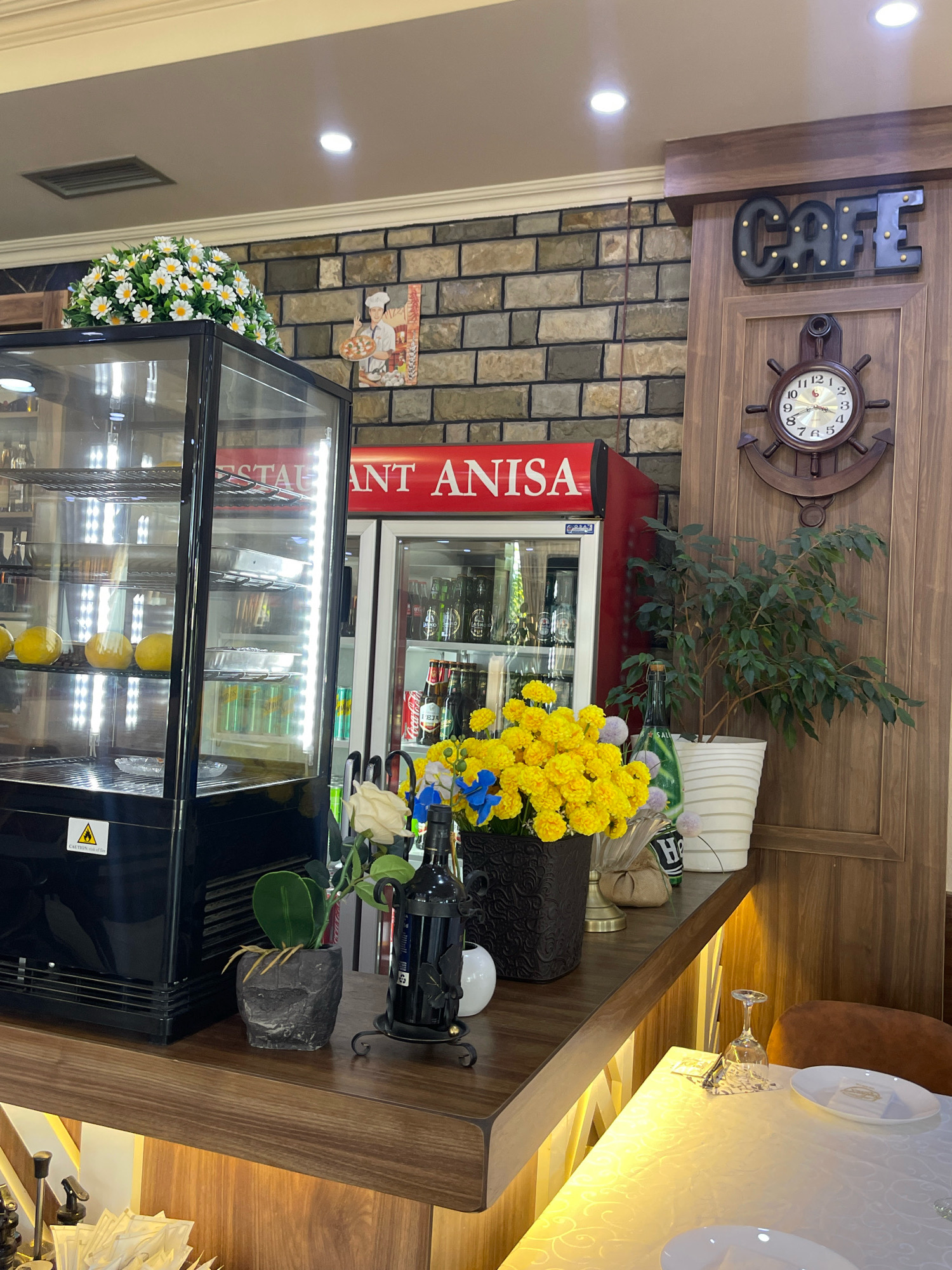The image depicts the interior of a cafe in Mirushë, Kosovo. The scene is dominated by a wooden counter and shelves, displaying various items. On the counter, a glass display case with a black frame holds several round, yellow objects, likely fruits. To the right of the display case, a red refrigerator with the "RESTAURANT ANISA" logo is visible. Further to the right, a potted plant with green leaves sits on a wooden shelf. Above the shelf, a decorative clock in the shape of a ship's wheel is mounted on a wooden panel. A dark sign with illuminated letters spelling "CAFE" is positioned above the clock. A bouquet of yellow flowers is placed in a dark pot on the counter, with a white rose and a decorative bottle nearby. In the background, a brick wall serves as a backdrop for some of the displayed items. A small, framed picture of a chef holding a pizza is hung on the brick wall. The table in the foreground is covered with a white tablecloth, and white plates and a wine glass are set on it. The lighting appears to be a mix of artificial lights from ceiling fixtures and display case lighting, creating a warm ambiance. The overall impression is that of a cozy and well-decorated cafe.