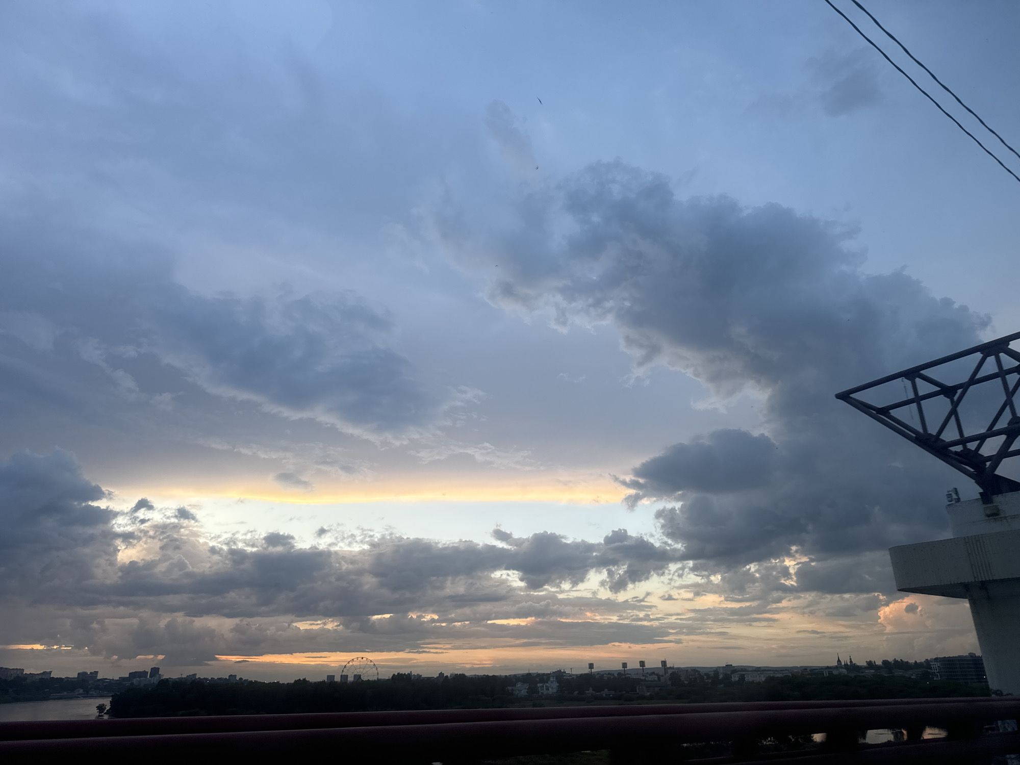 The image captures a dramatic sky over Irkutsk, Russia, likely during twilight or just after sunset. The sky is filled with a multitude of clouds, ranging from light gray to dark, ominous hues, with patches of warm orange and yellow light breaking through near the horizon, suggesting the last remnants of sunlight.
Dominating the right side of the frame is a large, angular, dark metal structure, possibly part of a bridge or industrial equipment, with a white concrete support. In the distance, a silhouetted cityscape unfolds, featuring buildings and what appears to be a Ferris wheel. A body of water, likely the Angara River, is visible in the foreground, with a dark railing obscuring the immediate view of the water. The overall atmosphere is one of natural grandeur and the encroaching darkness of evening.