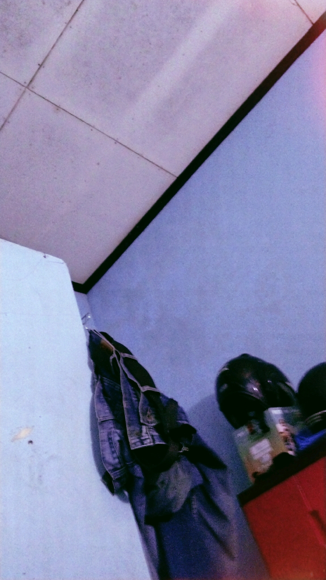 The image captures a dimly lit corner of a room, likely in Bergas, Indonesia. The scene is dominated by a light blue wall and a pale ceiling composed of tiled panels. On the left side of the frame, a white wall meets the blue wall at a corner. Hanging on a hook on the blue wall is a dark blue denim jacket over a pair of dark pants. To the right, on a red surface, two black helmets are visible. One helmet is more clearly in focus, and a colorful object, possibly a box or a book, is placed beside it. The overall impression is one of a domestic or personal space, possibly a bedroom or a storage area, with clothing and personal protective gear suggesting a resident who might ride a motorcycle. The lighting is low, and a film-like graininess is present throughout the image.