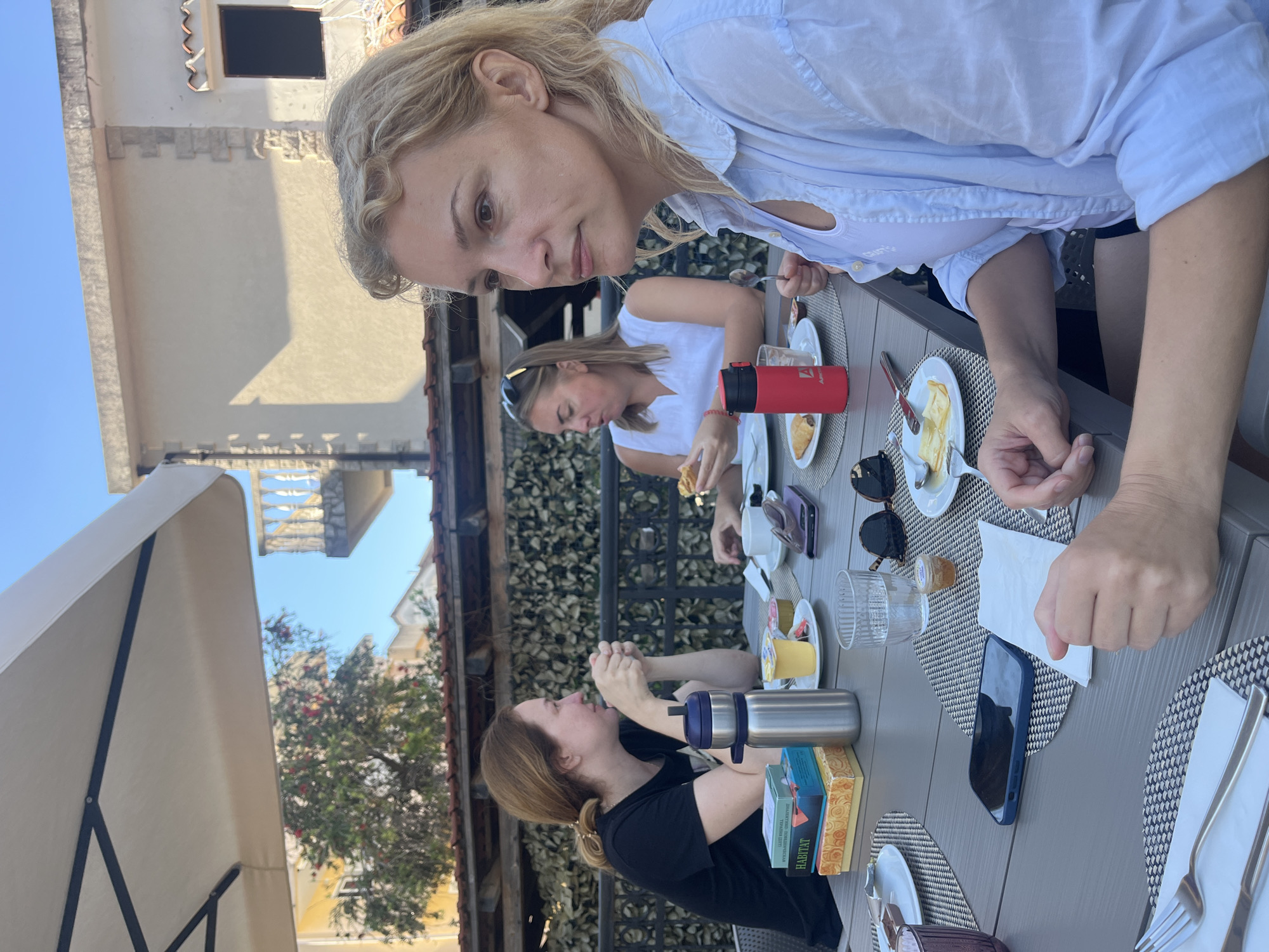 The image depicts three adult women seated at a grey outdoor dining table under a large, beige rectangular patio umbrella, in what appears to be a terrace or outdoor dining area in Budva, Montenegro. The setting is daytime under a clear blue sky, with bright sunlight casting shadows. In the foreground on the right, a blonde woman, approximately 40-50 years old, with wavy hair, is shown in profile. She wears a light blue button-up shirt over a white tank top. Her right arm rests on the table, and her left hand holds a spoon. She gazes towards the left side of the frame with a neutral expression. In the mid-ground, centered at the table, another woman, approximately 30-40 years old, with light brown hair pulled back, is depicted. She wears a white sleeveless top and has sunglasses perched on her head. She is actively eating, holding a pastry or similar food item with both hands near her mouth. To the left, in the background, a third woman, approximately 30-40 years old, with brown hair tied in a ponytail, is seated. She wears a black t-shirt and has her hands clasped on the table in front of her, looking towards the center woman. The table holds multiple items indicating a meal. From left to right: a stack of items including a box labeled "HABITAT" and another with Cyrillic script; a tall silver metallic thermal bottle; a dark blue smartphone in a case, screen down; a folded white paper napkin; a clear glass containing water; a small container, possibly of jam or honey; dark sunglasses; various white plates with food remnants (including what appears to be a pastry and a slice of cheese or butter); a white teacup and saucer; a yellow plastic cup (possibly yogurt); and a red thermal bottle with a black cap and an 'A' logo. Each place setting has a placemat with a geometric grey and white pattern. The immediate background behind the women features a dark metal railing or fence, beyond which is a textured wall constructed from stacked wooden elements or a similar material, possibly serving as a privacy screen. Above this, a red-tiled roof is visible. Further in the background, several light-colored stucco buildings with tiled roofs are present. One building on the far right has a dark, rectangular window opening, and another building exhibits a white stone balustrade on a balcony. Green foliage with