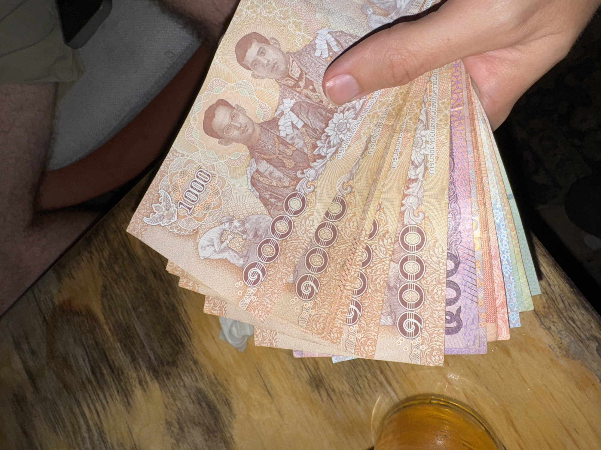 A hand is holding a fan of Thai Baht banknotes. The banknotes are predominantly orange and pink, with some purple and blue visible on the lower denominations. The top banknote features the number "1000" and portraits of two men in traditional attire. The banknotes are fanned out on a wooden surface, possibly a table. A glass with a yellow liquid, likely beer or a similar beverage, is visible on the left side of the image. In the background, parts of a person's legs and torso are visible, with a phone lying on a grey surface. The overall lighting suggests an indoor, possibly dimly lit setting.