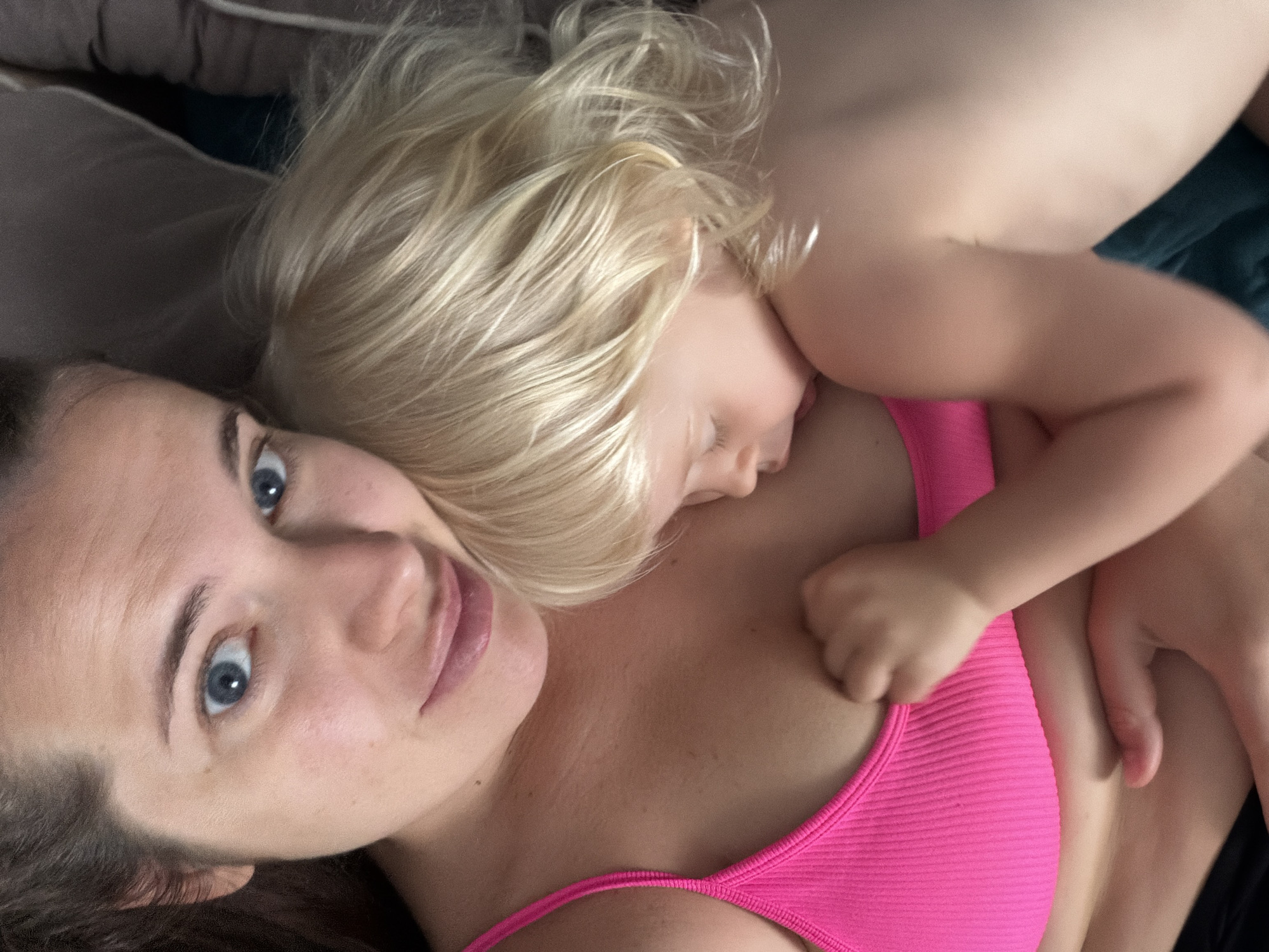 The media file shows a close-up, overhead view of a woman and a child lying down. The woman has brown hair and blue eyes, and is looking directly at the camera with a slight smile. The child, who is blond and appears to be very young, is nestled against the woman's chest, with their face turned away from the camera and their eyes closed. The child's arm is around the woman's neck. Both individuals are wearing comfortable clothing suitable for resting or sleeping. The woman is wearing a pink ribbed tank top. The background is soft and out of focus, suggesting an indoor setting, possibly a bed or couch. The lighting appears natural, indicating it could be daytime. The overall mood is intimate and peaceful, conveying a sense of closeness and comfort between the two subjects. The location context is Sochi, Russia, though no specific visual cues from Sochi are apparent in the image itself.