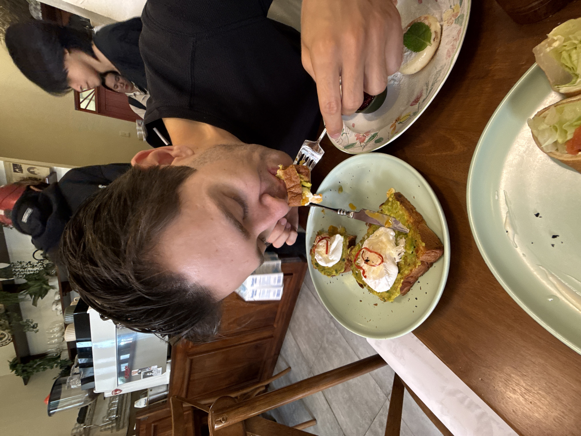 The image depicts an indoor scene at what appears to be a cafe or restaurant, likely during daytime hours. In the foreground, a man with short, dark hair and stubble is captured in the act of eating. He is wearing a dark long-sleeved top and is using a fork and knife to bring a bite of food to his open mouth, his eyes focused downwards.
On the wooden table in front of him, several food items are present. A light green plate holds two servings of toast topped with mashed avocado or guacamole and poached eggs, garnished with a red sauce or oil and black seeds. Another plate with a floral pattern contains a small pastry, possibly a pancake or scone, dusted with powdered sugar and adorned with a mint leaf, next to a small dish of red jam or preserve. A partially visible second light green plate shows two wraps or rolls filled with lettuce and other ingredients.
In the background, other people are seated at tables. One person with dark hair and a dark top is visible to the left, while another individual with dark hair is seen further back, observing the main subject. The cafe's interior features wooden tables and chairs, glass display cases, and what appears to be a counter area, illuminated by natural light entering through a window. The overall atmosphere is casual and relaxed. There is no clearly legible text visible within the frame. The setting is consistent with a modern cafe environment, such as one might find in Nha Trang Ward, Vietnam.