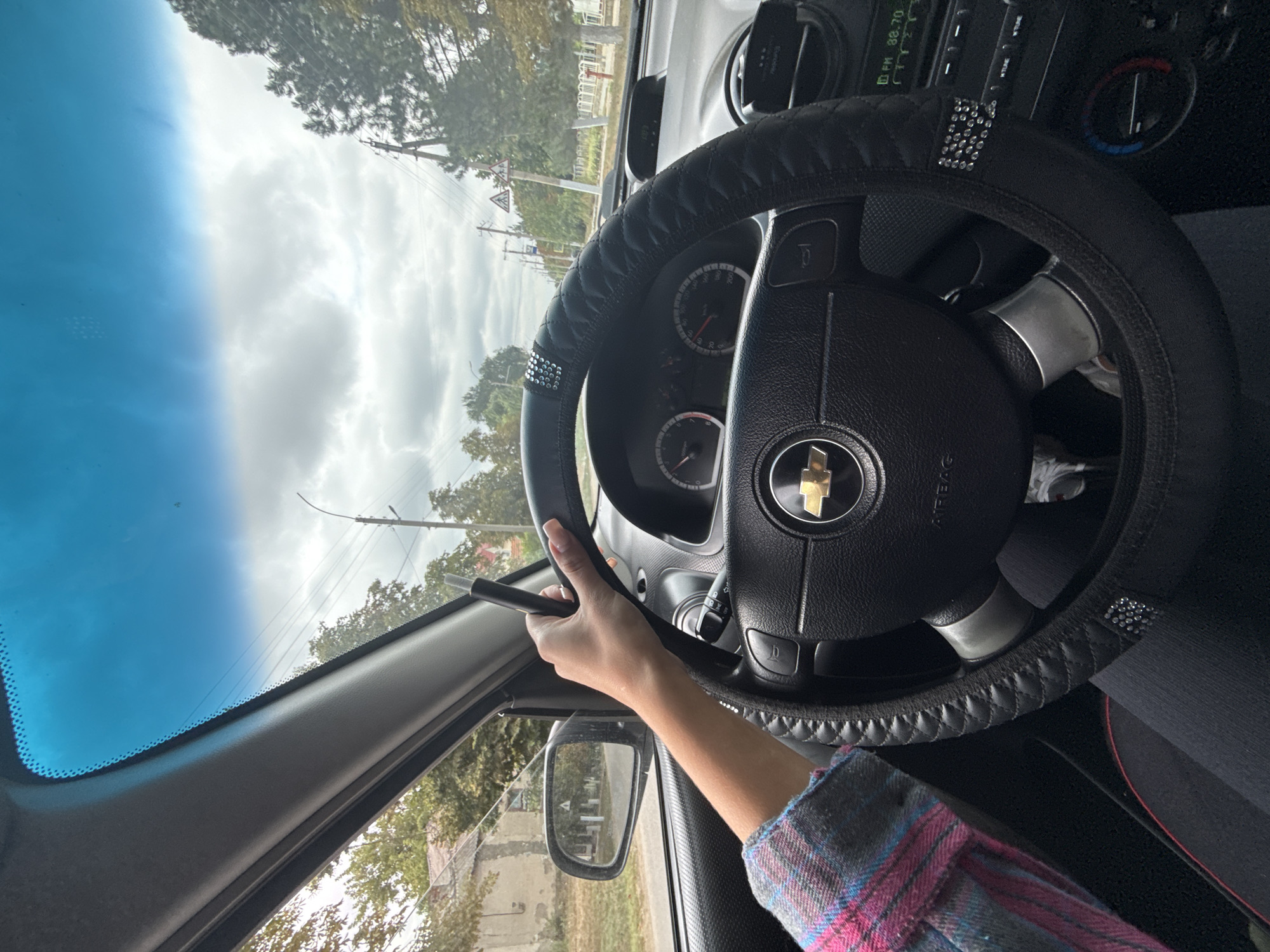 The image is a first-person perspective shot from inside a car, looking forward through the windshield. A woman's left hand, with manicured nails, is holding a black electronic cigarette device. Her arm is visible, wearing a pink, blue, and grey plaid long-sleeved shirt. The car's steering wheel is in the foreground, covered with a black quilted steering wheel cover adorned with rhinestones. The dashboard displays the car's instruments and radio. The car is on a paved road in Basarabeasca, Moldova. To the left, a rearview mirror reflects a building and trees. Outside the car, the environment appears semi-urban with houses, trees, and utility poles visible along the roadside. The sky is overcast with clouds.