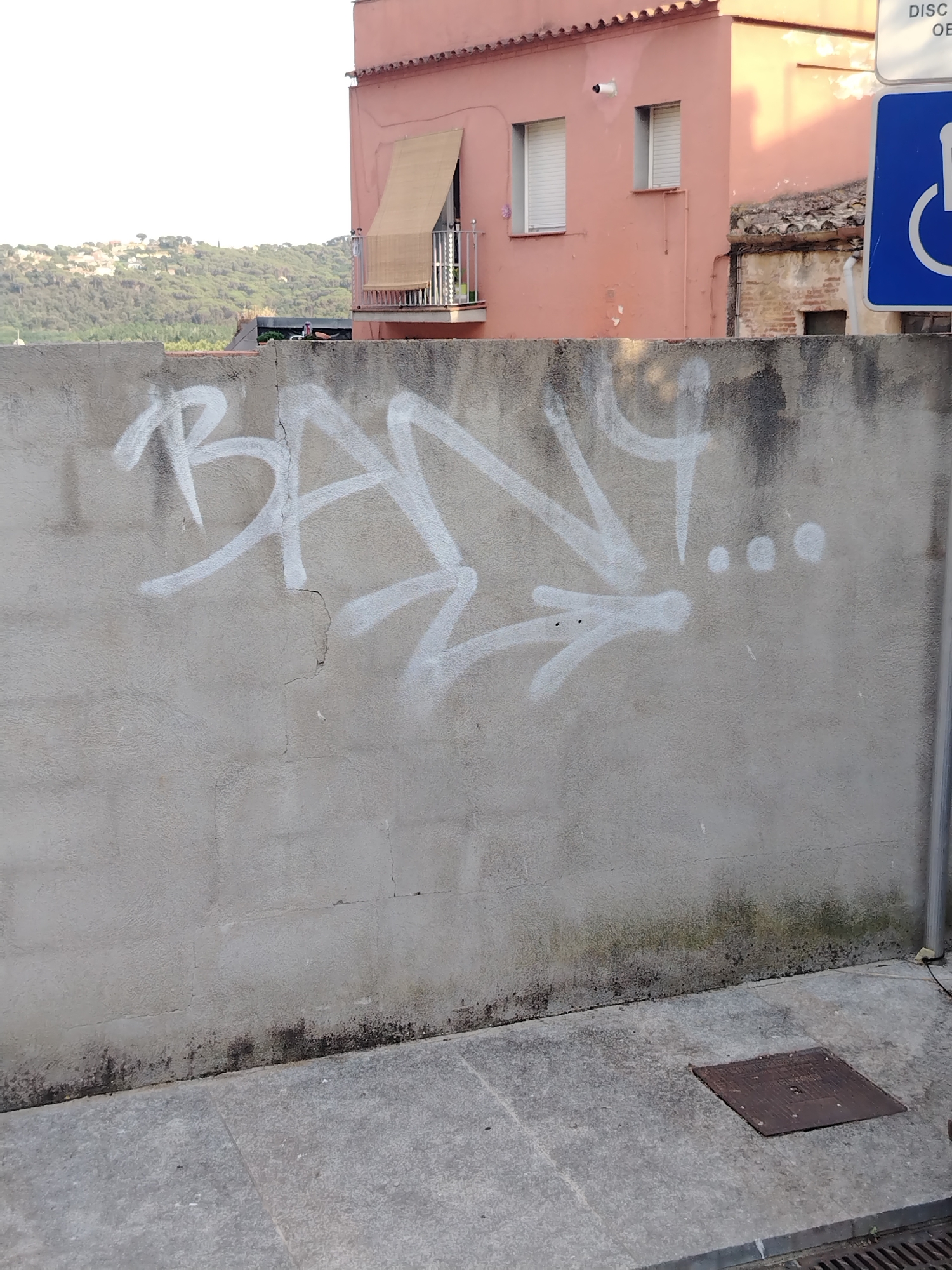 The image shows a graffiti-covered concrete wall in Hostalric, Spain. The graffiti, rendered in white spray paint, features stylized lettering that appears to read "BANY" followed by a series of dots. Behind the wall, a building with a pink facade and brown tiled roof is visible, with windows and a balcony. Further in the background, a lush green hillside dotted with houses can be seen under a bright sky. To the right of the building, a blue sign with a white wheelchair symbol indicates a disabled parking space. The foreground consists of a paved sidewalk and a metal drain cover. The scene captures a segment of an urban environment with a focus on the graffiti art.