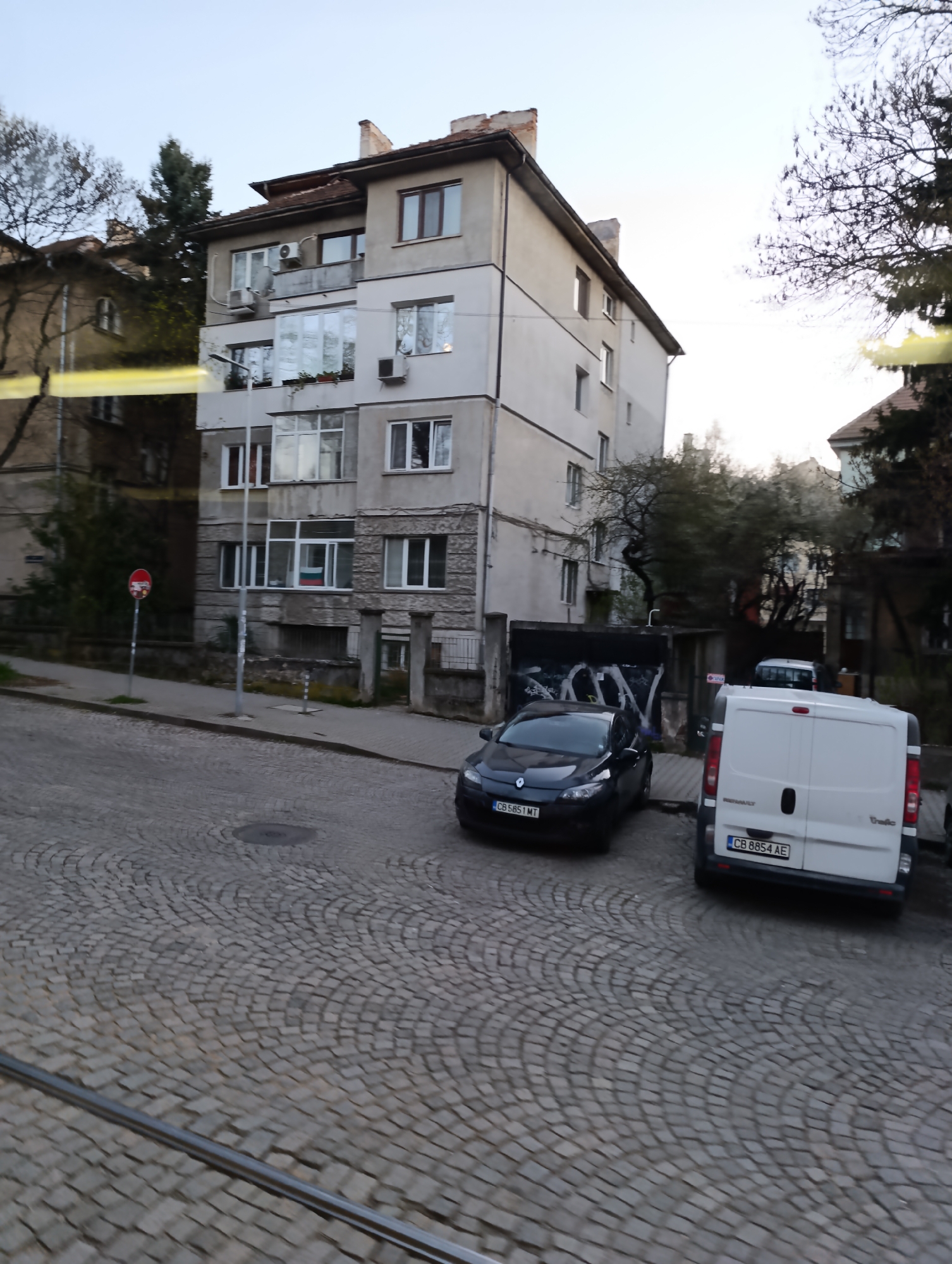 The image depicts a street scene in Sofia, Bulgaria, characterized by cobblestone paving and buildings of early to mid-20th-century architecture. A grey, multi-story residential building with balconies and window boxes is prominent on the left. Parked on the street are two vehicles: a dark-colored car (Renault Megane) with Bulgarian license plate CB 5851 MT and a white van (Renault Trafic) with license plate CB 8854 AE. A red and white traffic sign is visible near the building. Overhead tramway tracks run along the street. The weather appears overcast, with trees partially obscuring other buildings in the background. The general condition suggests an established urban neighborhood.