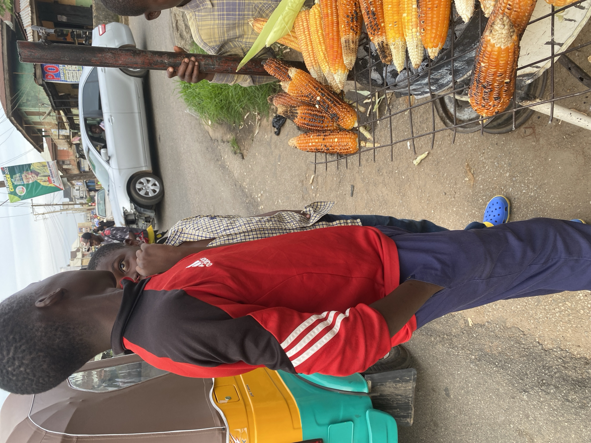 A young Black male stands in the foreground, facing away from the camera with his right hand in his pocket. He is wearing a red and black long-sleeved shirt with white Adidas stripes on the sleeves, and navy blue pants. To his left, a partially visible yellow and teal motorized tricycle is parked. Behind the young man, another boy in a plaid shirt is partially visible, looking upwards.
In the middle ground, a street scene unfolds. A silver sedan, which appears to have sustained front-end damage, is driving on the road. On the right side of the road, a charcoal grill is loaded with numerous ears of roasted corn. A person, likely an adult, is partially visible behind the grill, holding a metal rod. The background features a row of buildings and an overcast sky. A campaign poster with an image of a man and text, including "THE PEOPLE'S CHOICE 2023," is visible on a pole. The scene depicts a street-level view in a city, possibly Jos, Nigeria, characterized by street vendors, vehicles, and local architecture. The ground is asphalt and dirt.