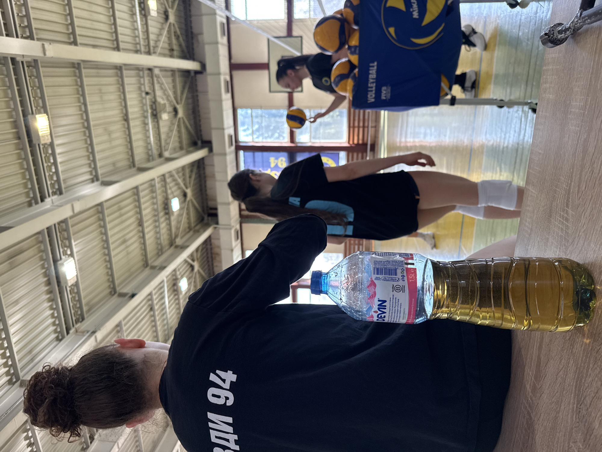 The image depicts an indoor volleyball training scene in Sofia, Bulgaria. There are three female volleyball players. One is close to the camera, viewed from behind, wearing a black t-shirt with "ВДИ 94" printed on it. A partially filled plastic water bottle labeled "DEVIN" is on the table in front of her. Another player is in the middle ground, wearing a black top and white knee pads, practicing volleyball skills. The third player, also in a black top, is in the background, with a volleyball. A blue volleyball cart filled with balls is also visible. The scene is set inside a volleyball gymnasium, evidenced by the volleyball court and ceiling structure.