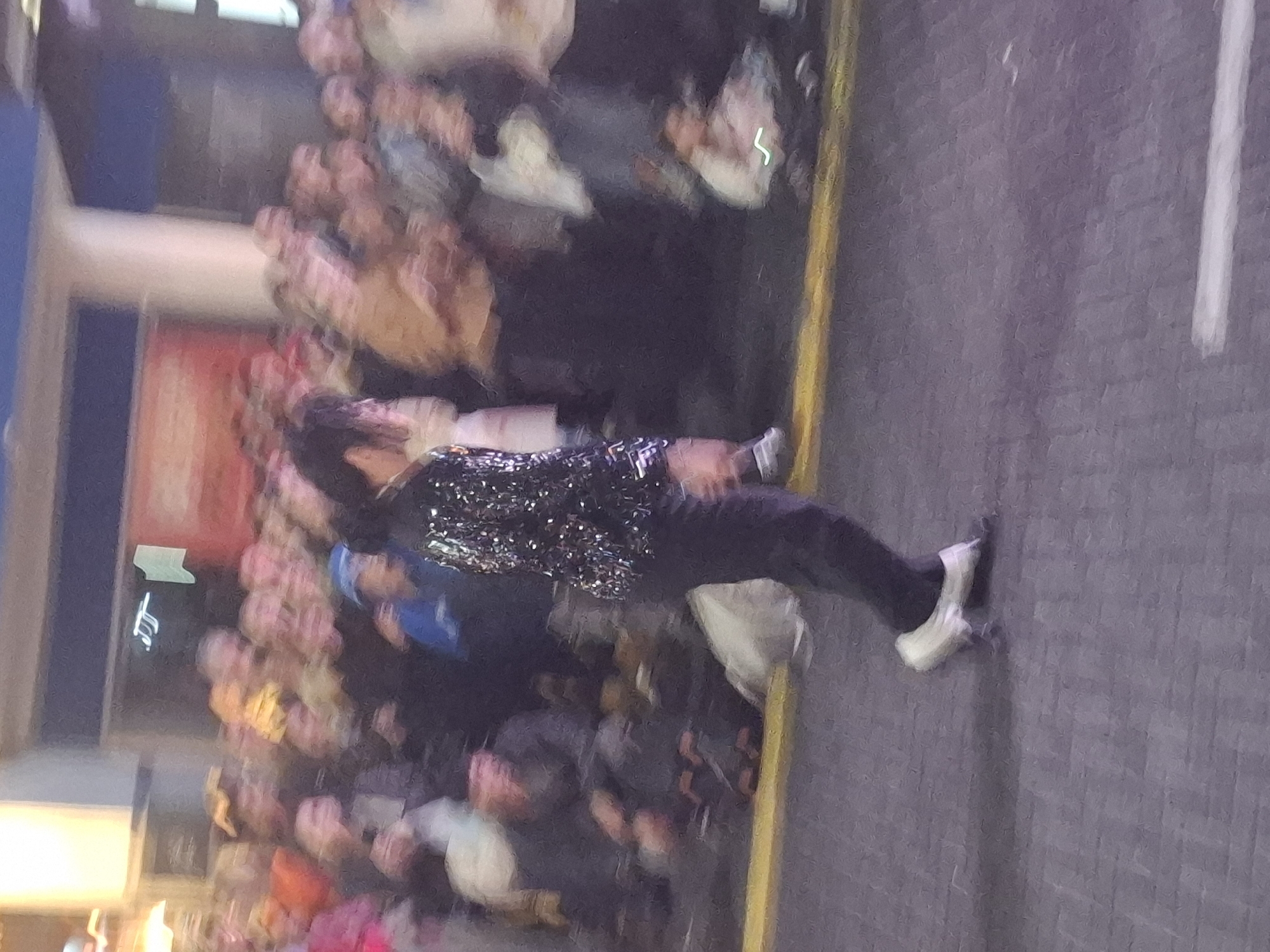 A person in a sparkling top and dark pants stands on a street, interacting with a crowd of people blurred in the background. The setting appears to be an urban environment, potentially in Buenos Aires, Argentina. A white line is visible in the lower right corner, suggesting a street marking. A building is visible behind the crowd, with some storefront details.