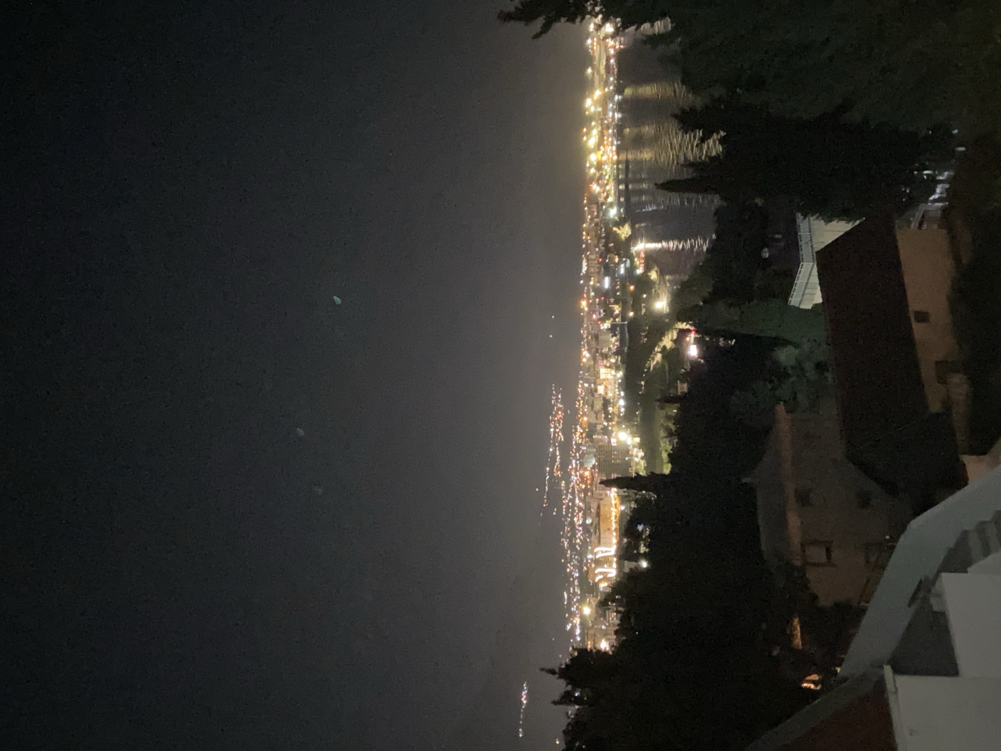 A night view of Šušanj, Montenegro, shows a densely populated coastal area with numerous lights illuminating the landscape. Buildings are visible, interspersed with trees and vegetation. The city stretches along the coastline, reflecting light into the dark water. In the foreground are architectural structures that include buildings, possibly residences, or hotels. The sky is dark and clear, suggesting a calm night.