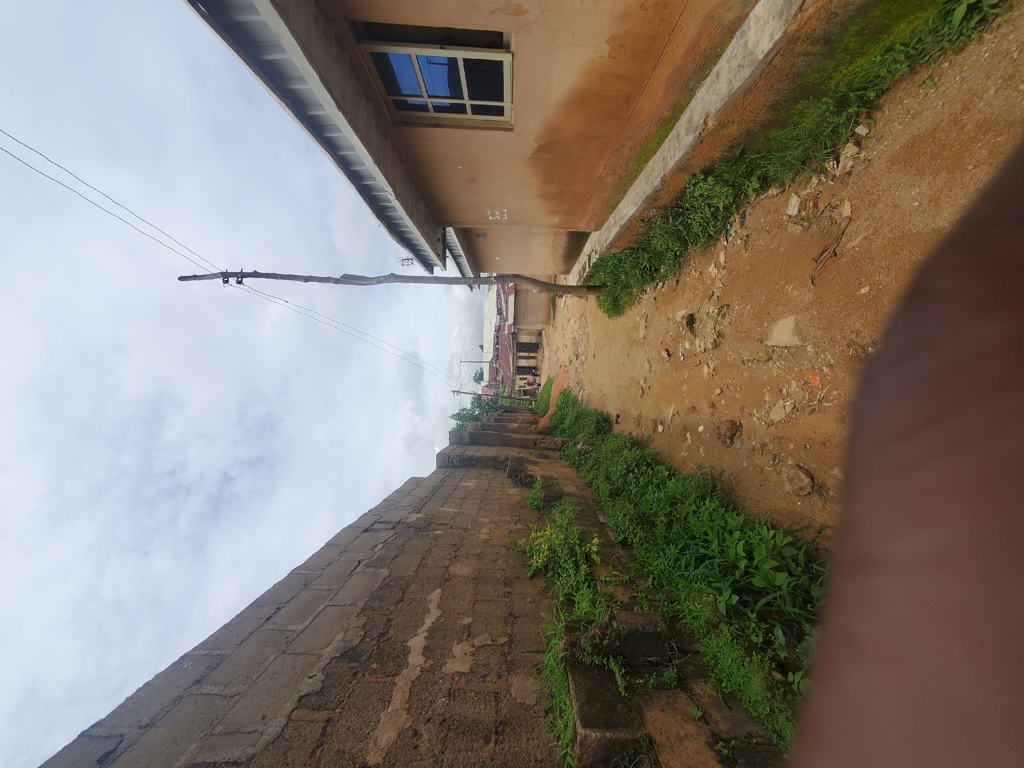The image depicts a narrow, unpaved alleyway in an urban environment. On the left side of the alley is a tall, textured concrete block wall, partially covered with green vegetation. On the right side, a building with light brown, weathered walls and a grey, corrugated roofline stands. A rectangular window with blue-tinted panes is visible on this building. A thin, rustic wooden pole with electrical insulators and wires stretches upwards from the ground in the middle of the alley. The ground of the alley is composed of dirt, gravel, and scattered debris. In the distance, more buildings and structures are visible, suggesting a residential or commercial area. The sky is overcast with clouds. A portion of a person's hand or arm is visible in the foreground, obscuring the lower part of the scene.