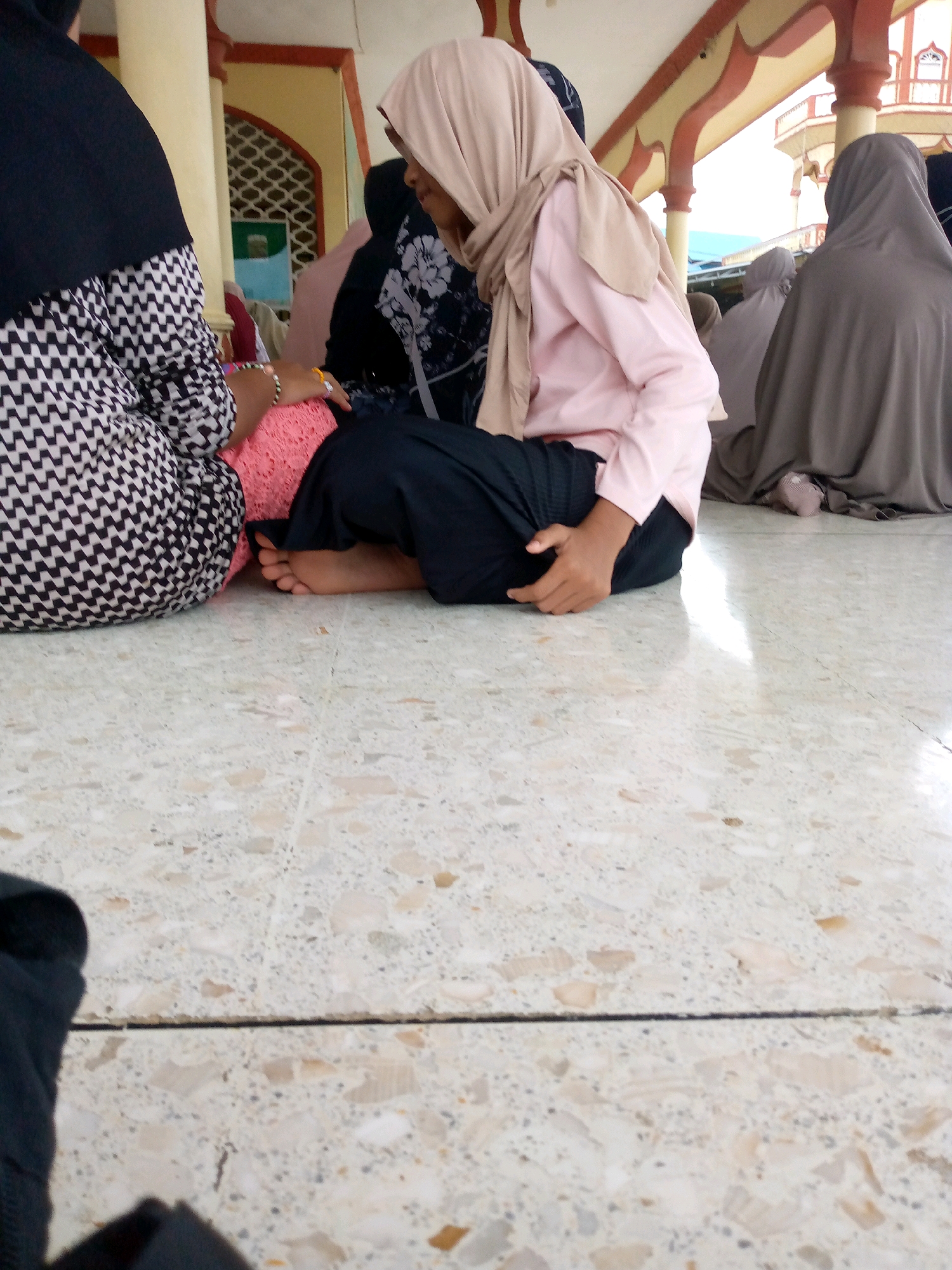 In Keudeteupinpunti, Indonesia, a group of women are seen sitting on a terrazzo floor. One woman in the foreground is wearing a light pink hijab and a light pink long-sleeved shirt, with black pants. She is resting her left hand on her thigh and appears to be holding a pink cushion with her right hand. Next to her, another woman is visible from the waist down, wearing a black and white patterned top and black pants. Behind them, several other women are also seated, their faces obscured by their hijabs, which are in various shades of gray and taupe. The background shows architectural elements of what appears to be a building, with arched doorways and columns, suggesting a communal or religious gathering space. Sunlight creates bright reflections on the floor.