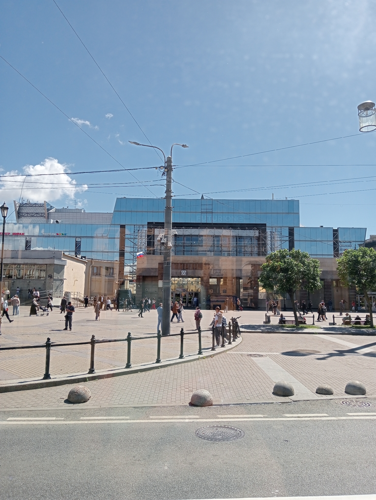 A sunny day in Saint Petersburg, Russia, is captured in this wide-angle shot of a public square. A modern glass-fronted building, likely a commercial or cultural center, dominates the background. Its reflective facade mirrors the clear blue sky and scattered white clouds, giving it a contemporary feel. In front of the building, a bustling plaza is filled with people engaged in various activities. Some are strolling, others are gathered in small groups, and a few are pushing strollers. The ground is paved with interlocking bricks, and a black metal railing separates the pedestrian area from a road in the foreground. Large, rounded concrete bollards are strategically placed to delineate the space.
A tall lamppost with multiple lights stands prominently on the right side of the frame, its wires stretching across the sky, indicating the presence of an urban infrastructure. A Russian flag is visible flying from the facade of the building, adding a patriotic element to the scene. The overall atmosphere appears lively and relaxed, suggesting a typical day in a vibrant urban environment. The presence of trees and benches indicates that this is a public space designed for both commerce and leisure. The image seems to have been taken in the afternoon, given the bright sunlight and the length of the shadows. No specific event is overtly taking place, but the scene conveys the general activity of daily life in the city. Some visible text includes "ТЕАТР МИРАЖ" and "FUTURION" on the building facade.