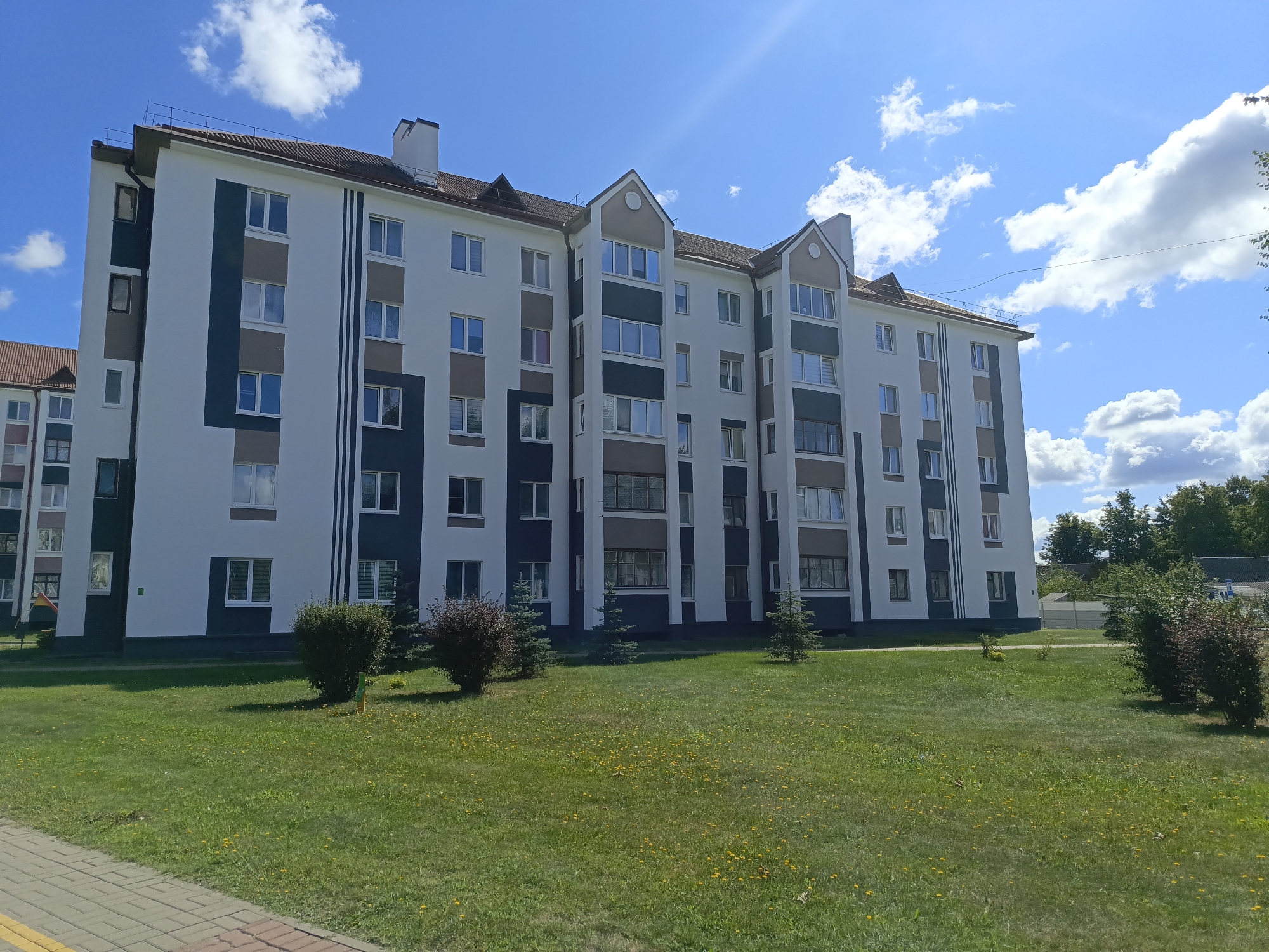 The image shows a modern, multi-story apartment building with a white facade accented with dark gray and brown panels. The building has a pitched, brown roof and numerous windows with white frames. In front of the building is a well-maintained lawn with scattered yellow dandelions and several small evergreen trees and bushes. A paved walkway is visible in the foreground on the left. The sky above is bright blue with scattered white clouds, and sunlight creates a lens flare in the upper right corner. The scene depicts a residential area in the city of Ivatsevichy, Belarus, on a sunny day.