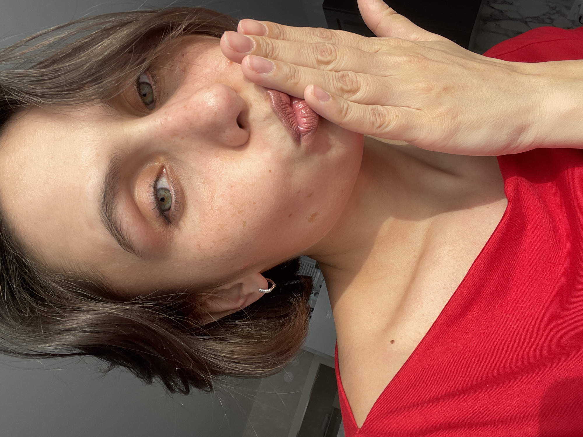 A woman with fair skin, freckles, and short brown hair poses for a selfie-style photo. She is wearing a red top. Her left hand is raised to her face, obscuring part of her cheek and mouth. She has light green eyes. She has a small earring in her left ear. The background is out of focus, but suggests an interior space. There is no visual information in the image to determine any location in Izhevsk, Russia.
