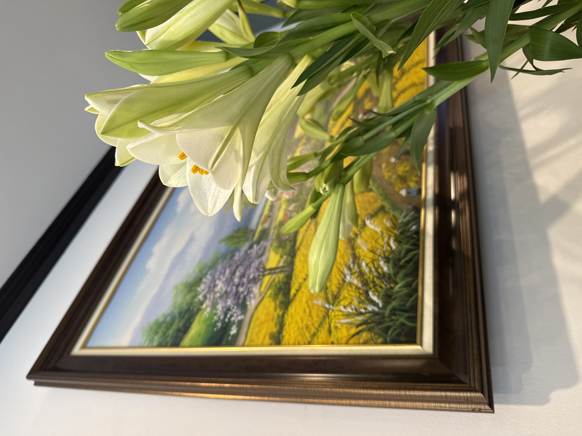 A close-up, angled shot shows a painting hanging on a white wall, with lilies in the foreground. The painting depicts a landscape with a blue sky, green fields and trees, and a large area of yellow flowers. A path winds through the scene. A white wall is partially visible behind the painting. A dark-colored molding is on the left wall. The lilies are mostly white with green buds and stems. The specific location within Hà Nội, Vietnam, is not discernible from the image.