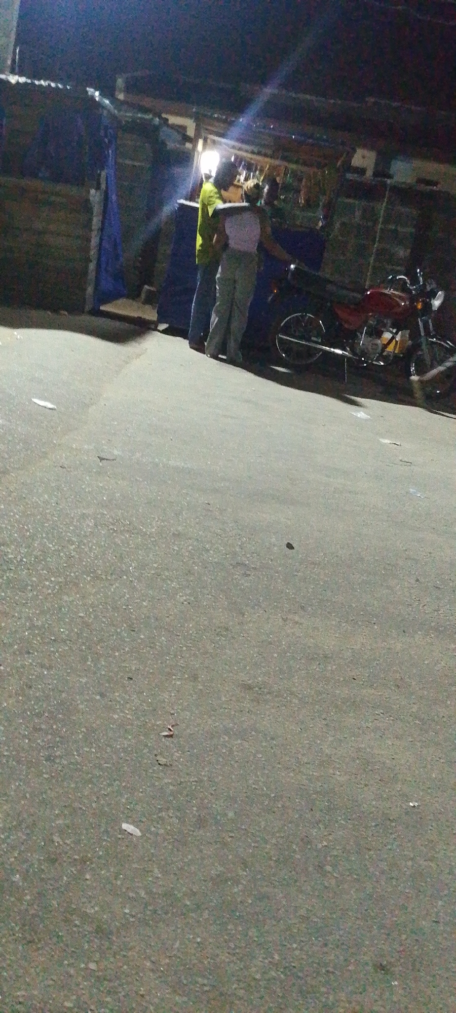 A dimly lit street scene, likely in Lokoja, Nigeria, depicts two individuals standing near a parked red motorcycle. The person on the left is wearing a green shirt and dark pants, while the person on the right is wearing a light top and pants. They are standing near what appears to be a market stall with blue tarpaulin coverings. Debris is scattered on the asphalt road. The scene appears to be at night, with limited artificial lighting creating strong shadows.