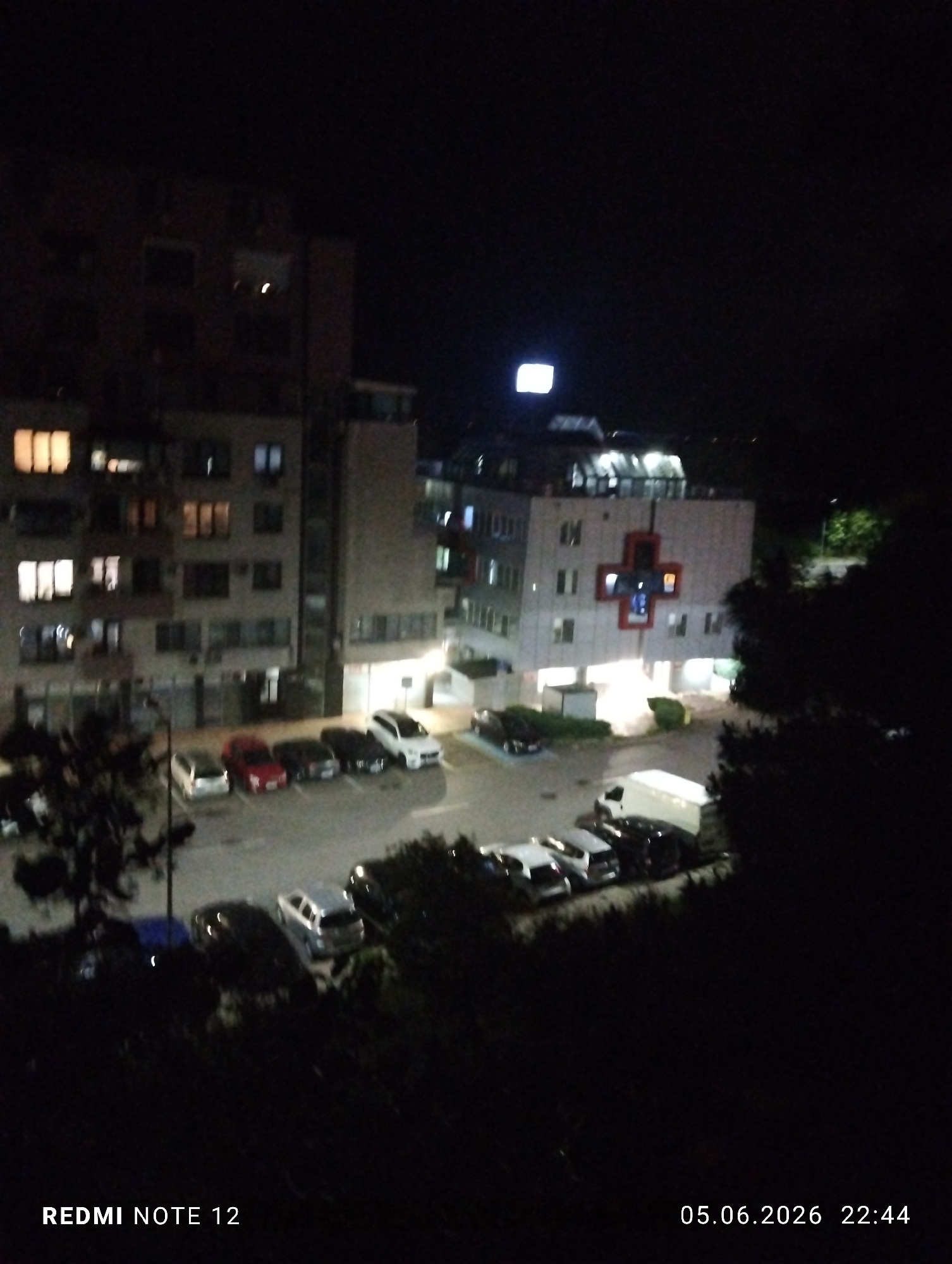 This forensic description details a nighttime scene captured in Burgas, Bulgaria, on June 5, 2026, at 22:44, using a Redmi Note 12. The image provides an elevated view of an urban environment.
The central and right portions of the image feature a prominent, multi-story building, identifiable as **ДКЦ 1 - Бургас ("Св. Климент Охридски" Polyclinic)**, due to a large, illuminated red cross emblem displayed on its facade. Above this building, a bright, rectangular light source is visible against the dark sky. To the left, another multi-story building, likely residential or mixed-use, displays numerous illuminated windows.
In the midground, a parking area is visible, accommodating multiple vehicles, including a red car, several dark-colored sedans, a silver minivan, and a white van. The ground surface appears wet, reflecting the artificial light sources from the buildings and streetlights, suggesting recent precipitation or a damp environment. Dark, indistinct foliage and trees partially obscure the lower foreground and the right periphery of the scene. No individuals are discernible within the frame. The overall lighting is low, consistent with a nighttime setting in an urban area.
