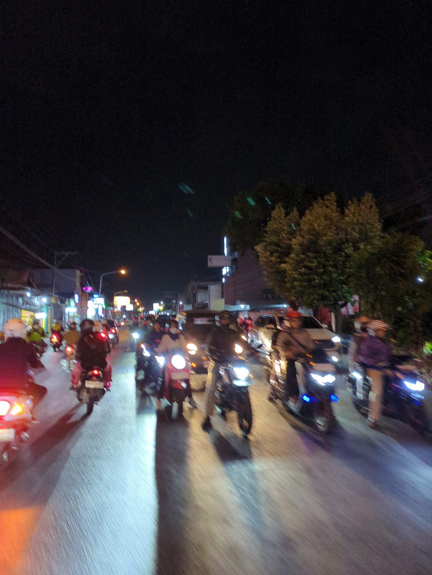 This image depicts a busy street scene at night in Gentan, Indonesia, characterized by numerous motorcycles and a dark sky. The perspective suggests the photographer is on a motorcycle, looking forward. Several motorcycles are visible in the frame, with their headlights illuminating the road. People are riding the motorcycles, many wearing helmets. The street is paved and appears to be well-lit by streetlights and the headlights of the vehicles. Buildings and trees line the side of the road, with some illuminated signs suggesting the presence of shops or businesses. The overall impression is one of traffic and activity on a street at night in a semi-urban or suburban setting.