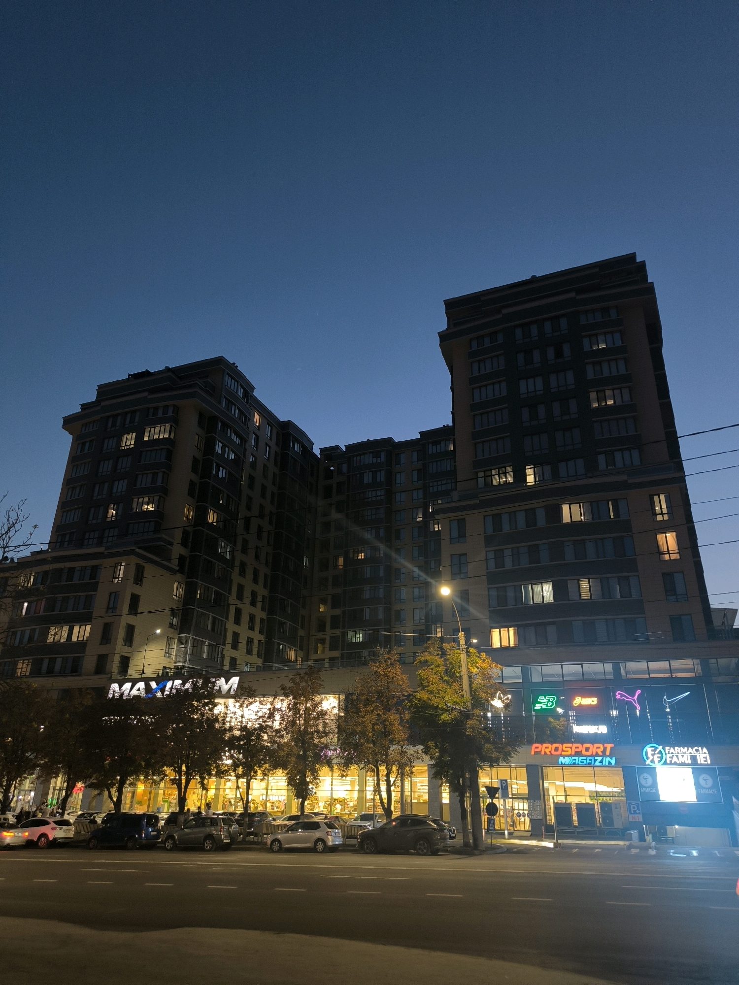 The image shows a modern, multi-story building at dusk in Chișinău, Moldova. The building features a mix of residential apartments and commercial spaces on the ground floor. Illuminated signs identify some of the businesses, including "MAXIMUM" and "PROSPORT MAGAZIN," as well as "FARMACIA FAMILIEI." The sky is a deep blue, transitioning into night. Several cars are parked along the street in front of the building, and streetlights illuminate the scene. Trees with autumn foliage are visible, adding a touch of natural color to the urban landscape. The architecture is contemporary, with numerous windows reflecting the interior lights. The overall atmosphere is one of a lively urban environment at the close of day.
