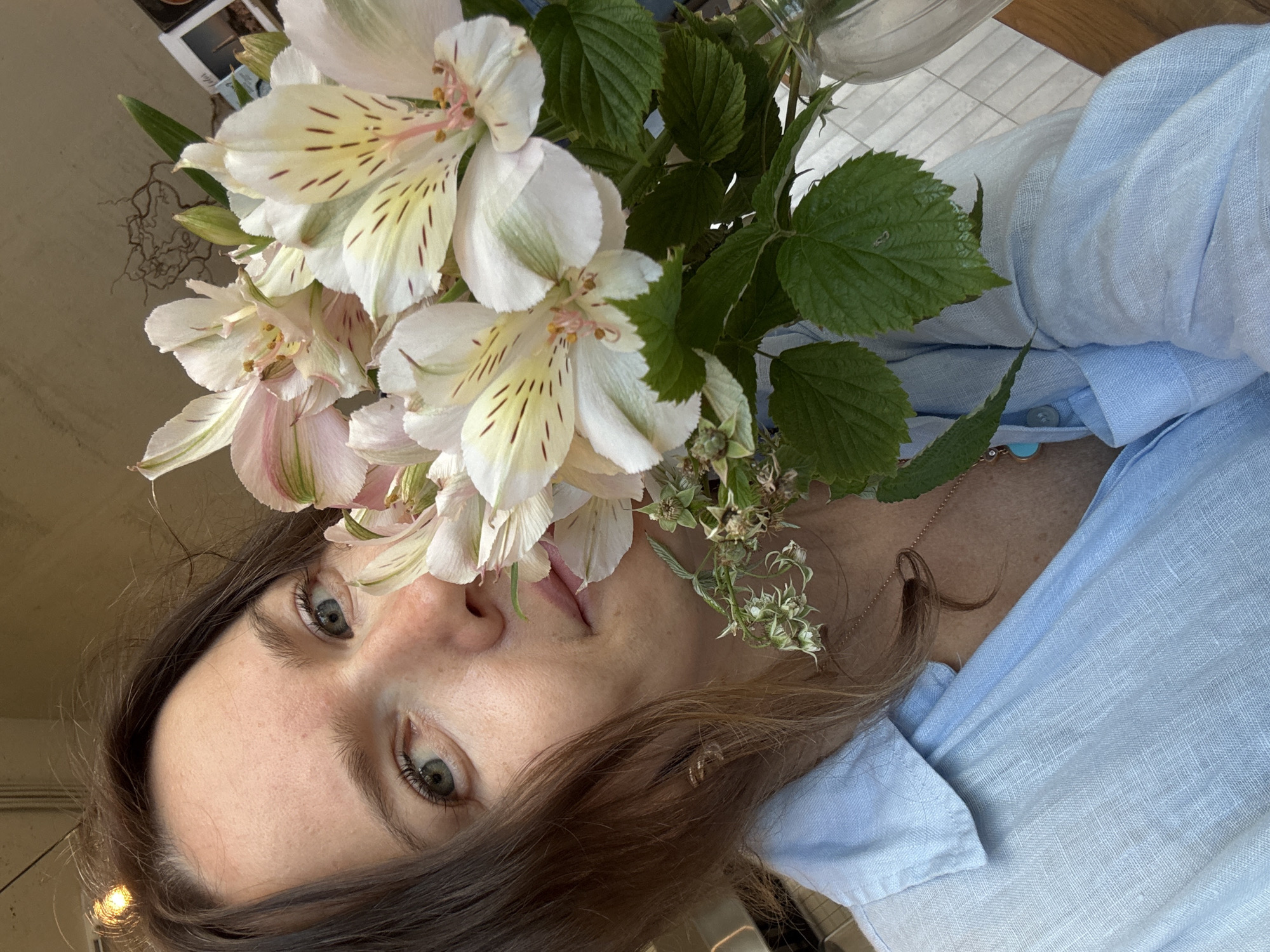A close-up, eye-level selfie shows a woman holding a bouquet of white and pink alstroemeria flowers, partially obscuring her face. She has brown hair and green eyes and is wearing a light blue linen shirt. A necklace with a turquoise pendant is visible. The background is blurred, but suggests an indoor setting with warm lighting. There is no indication of the city of Moscow, Russia.
