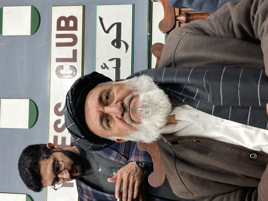 The image displays an indoor scene at the Quetta Press Club in Quetta City Tehsil, Pakistan, featuring two seated males. In the foreground, an elderly man with a full white beard, a black patterned turban, a brown jacket, and a white collared shirt beneath a striped black shawl, looks directly towards the viewer with a neutral expression. Behind him and slightly to his left, a younger man with dark hair, a beard, and eyeglasses is visible, wearing a plaid blazer over a dark shirt. This individual is looking downwards, holding a light-colored rectangular object, possibly a mobile phone, in his hands. A watch is visible on his left wrist. The background features a light grey wall with a prominent white sign bearing the English text "PRESS CLUB" in reddish-brown capital letters. Below this, another sign displays "كويئته" (Quetta) in Urdu script. Parts of a carved wooden chair back are visible behind the foreground subject.