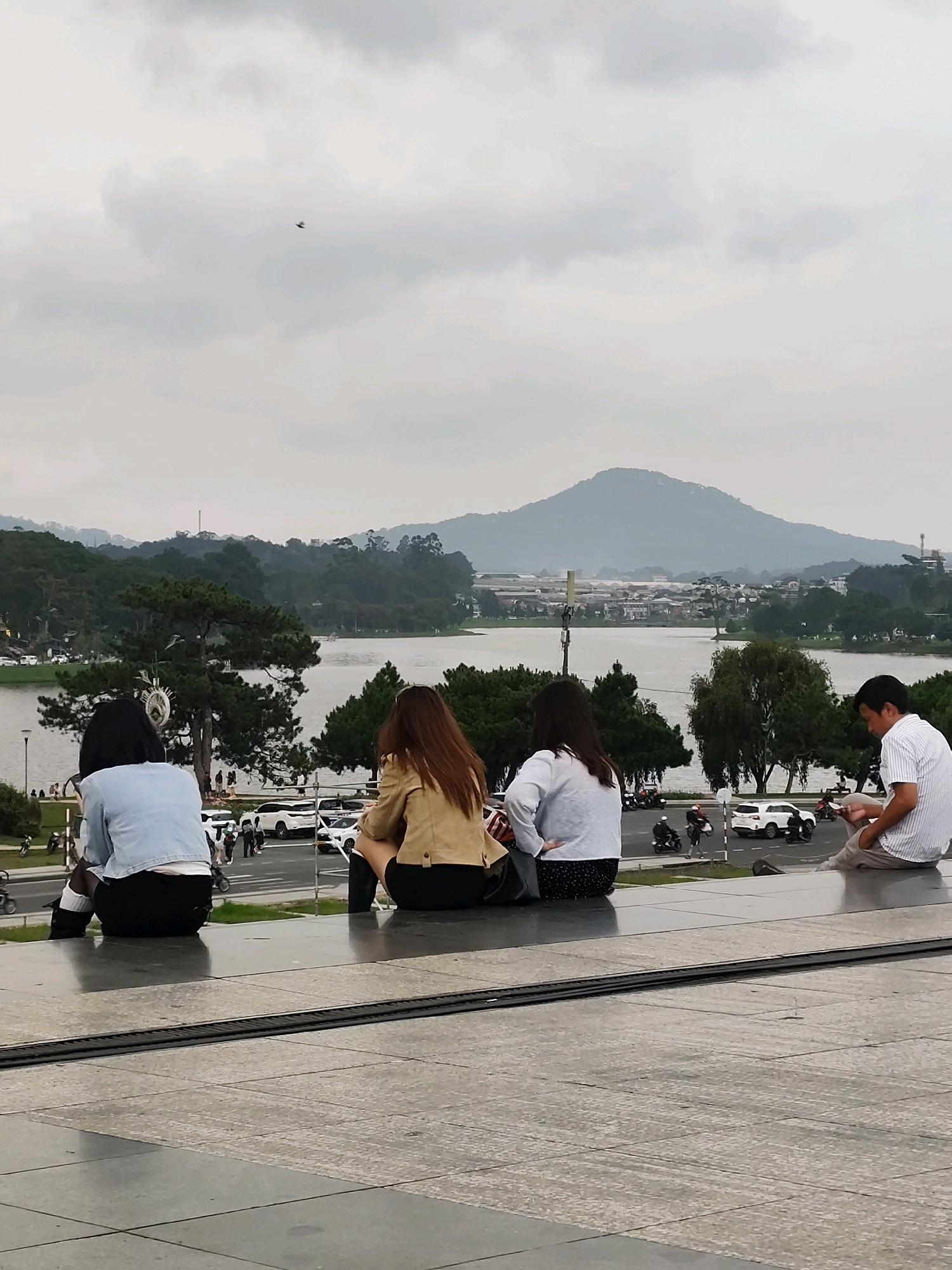 In the city of Da Lat, Vietnam, four individuals are seated on a raised concrete platform overlooking Xuan Huong Lake. Two women, viewed from behind, sit on the left side of the platform. One wears a light blue jacket and black shorts, while the other has on a tan jacket and black skirt. To their right, another woman with long brown hair, wearing a white sweater and black skirt, is also seated. On the far right, a man in a white pinstriped shirt and light-colored pants is seated, looking down at his phone.
The background features a tranquil lake surrounded by lush green trees and distant mountains, characteristic of Da Lat's scenic landscape. Buildings are visible on the far side of the lake, and a road with several cars and motorbikes is present between the platform and the lake. The sky is overcast with scattered clouds, and a single bird is visible in flight in the upper left quadrant of the image. The overall scene depicts a moment of quiet observation and relaxation in an urban park setting.