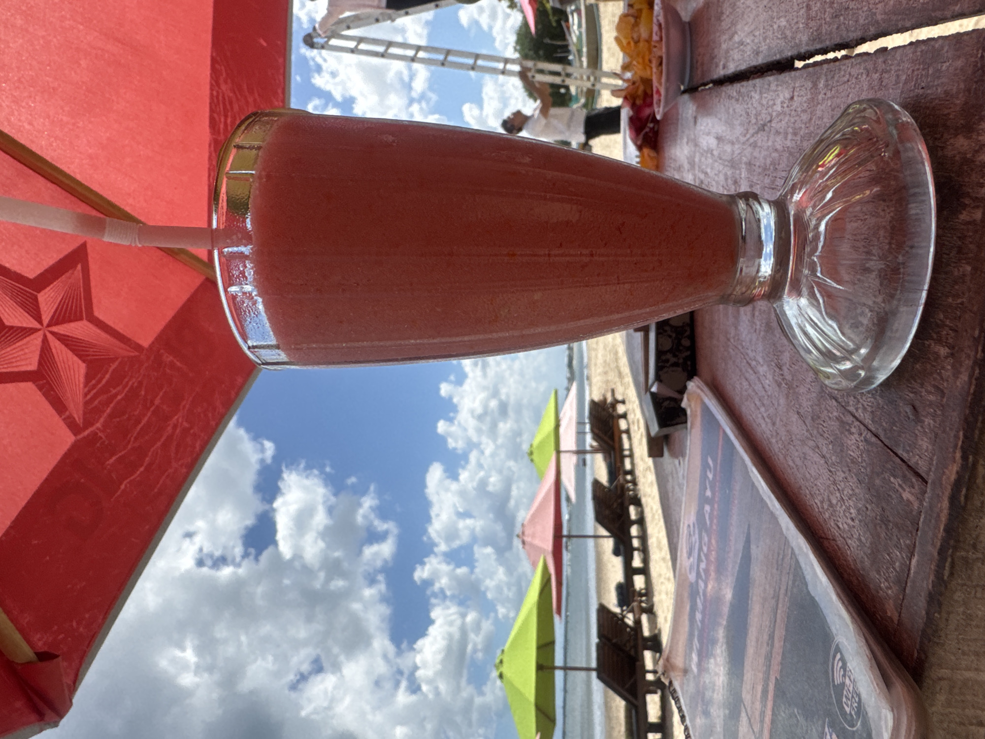 A tall, clear glass filled with a pink, blended beverage sits on a weathered wooden table in the foreground. A white straw extends from the drink, disappearing under the edge of a red umbrella. In the background, a sandy beach is visible with rows of empty lounge chairs and brightly colored umbrellas (green and pink). The ocean stretches out to the horizon under a bright blue sky dotted with white, fluffy clouds. On the right side of the image, two men are working on a tall ladder. One man, wearing a white shirt and dark pants, is near the top of the ladder, seemingly adjusting something. Another man, also in a white shirt, is standing on the ground, looking up at the ladder. A small plate with snacks is visible on the table to the right of the drink. A menu is lying on the table to the left of the drink, with the text "WI-FI FREE" visible. The scene suggests a relaxed, tropical setting, likely a beachfront cafe or restaurant in Kuta Selatan, Indonesia, on a sunny day.