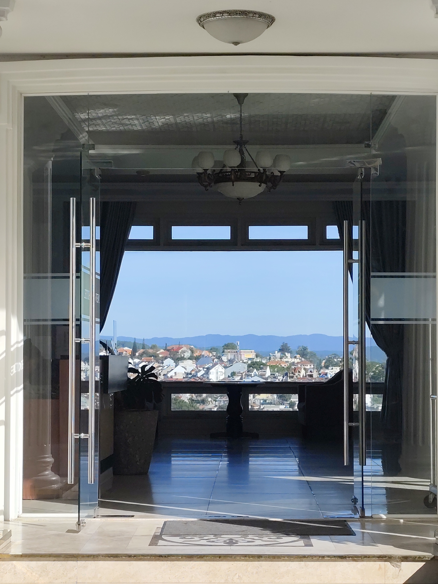 An open glass double doorway leads to a room with a view in Da Lat, Vietnam. The doorway is framed by white columns and the ceiling has a decorative light fixture. A large window frames a panoramic view of Da Lat, with rooftops and distant mountains visible under a clear blue sky. Inside the room is a table, potted plant, chandelier and curtains. The floor is tiled and reflective, and a doormat is visible at the threshold.
