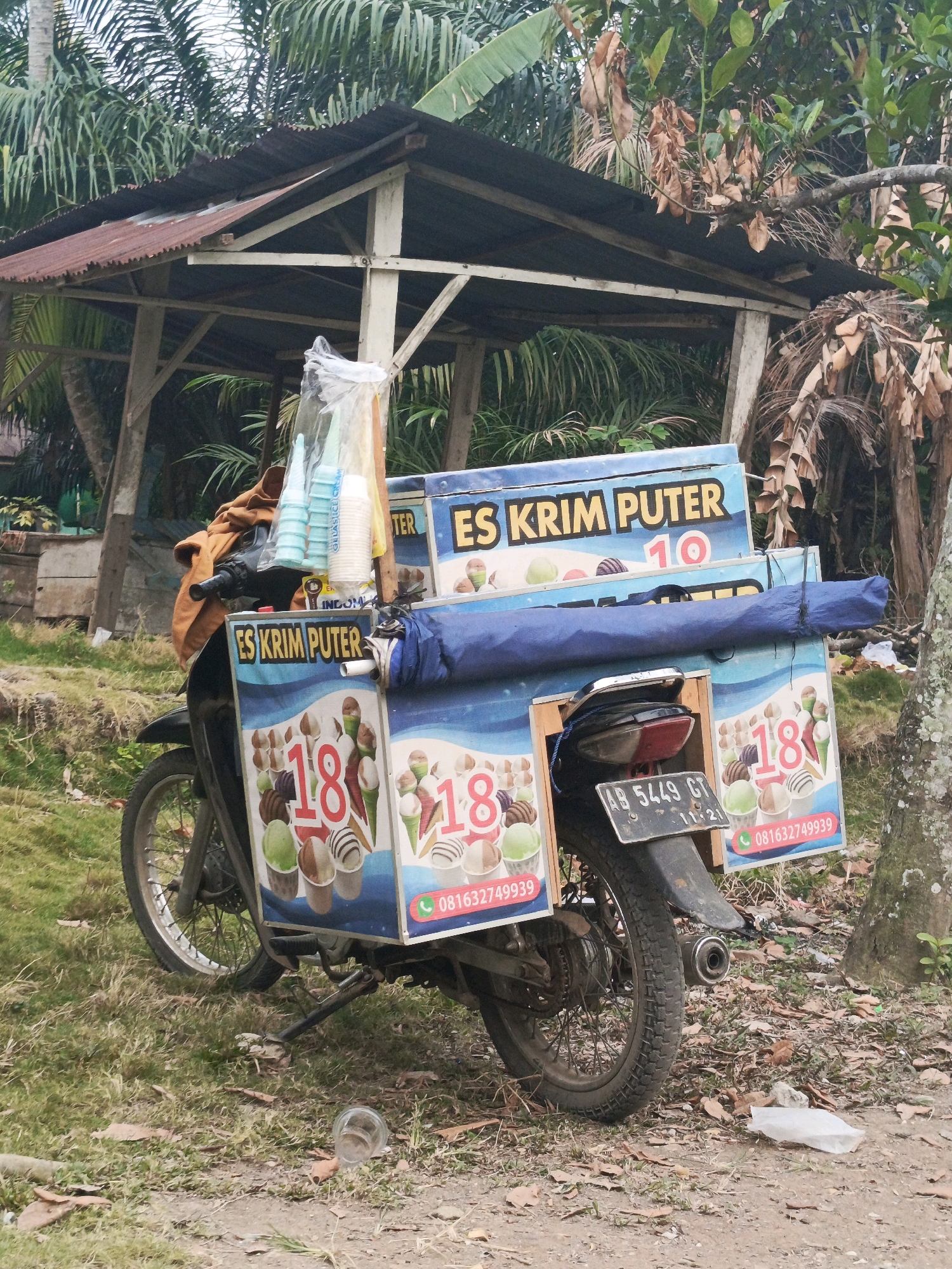 An ice cream cart, adapted from a motorcycle, is parked near a weathered, open-sided structure with a rusty tin roof. The cart, predominantly blue and white, displays advertisements for "ES KRIM PUTER" (rolled ice cream) and features images of ice cream cones. One side of the cart prominently features the number "18" in large red font, along with a phone number. Several plastic cups and cones are stacked and visible on the cart. The motorcycle itself is partially obscured by the cart, with its rear tire and license plate (AB 5449 GT 11-21) visible. The ground is covered in grass, dirt, and fallen leaves. Trees and foliage are present in the background. The scene appears to be set in an outdoor, somewhat unkempt area, possibly a roadside or a market stall location in Megang Sakti II, Indonesia. There are no people visible in the frame.