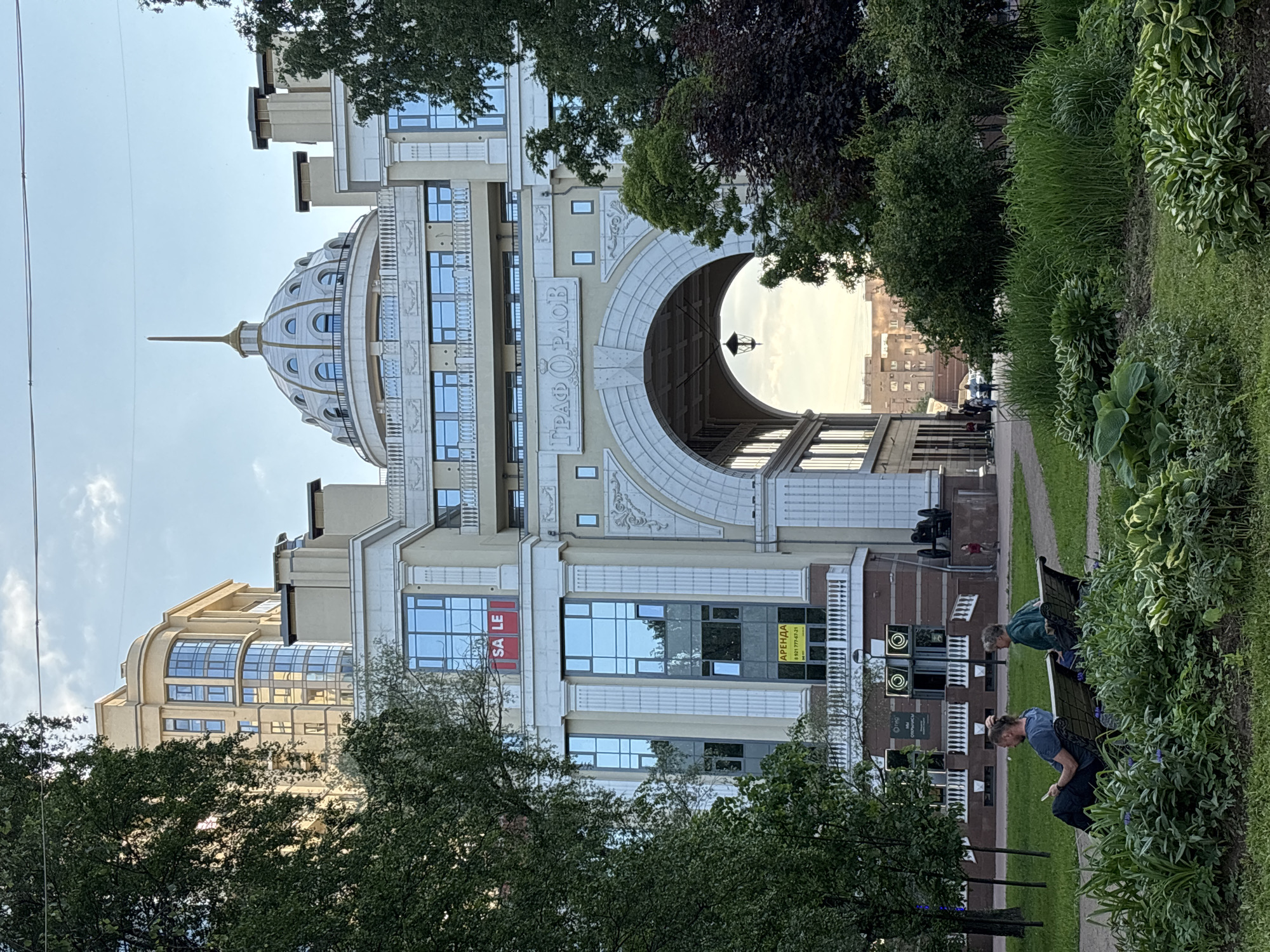 A grand, multi-story building with neoclassical and art deco architectural elements dominates the scene. Its light-colored facade features numerous windows, some arched and some rectangular, along with decorative mouldings and columns. A prominent central dome, topped with a slender spire, rises above the building. A large, arched pedestrian passage cuts through the lower section, within which a vintage-style hanging street lamp is visible. Above this archway, the Russian text "ГРАФ ОРЛОВ" (Count Orlov) is inscribed.
To the right of the building, a lush green strip of vegetation, including various bushes, hostas, and grasses, borders a paved pathway. Two individuals are seated on separate dark benches along this path, facing away from the camera. The person closer to the foreground appears to be engrossed in a device.
Visible on the building's windows are commercial signs: a red sign reading "SALE" and a yellow sign with "АРЕНДА" (RENT or FOR LEASE) and the phone number "8 921 777-63-21". Another section of the building on the far left features distinctive curved, multi-pane windows.
The lighting suggests late afternoon or early evening under clear skies, indicating fair weather. The scene is an urban setting, likely a park or promenade, adjacent to this ornate building in Saint Petersburg, Russia.
