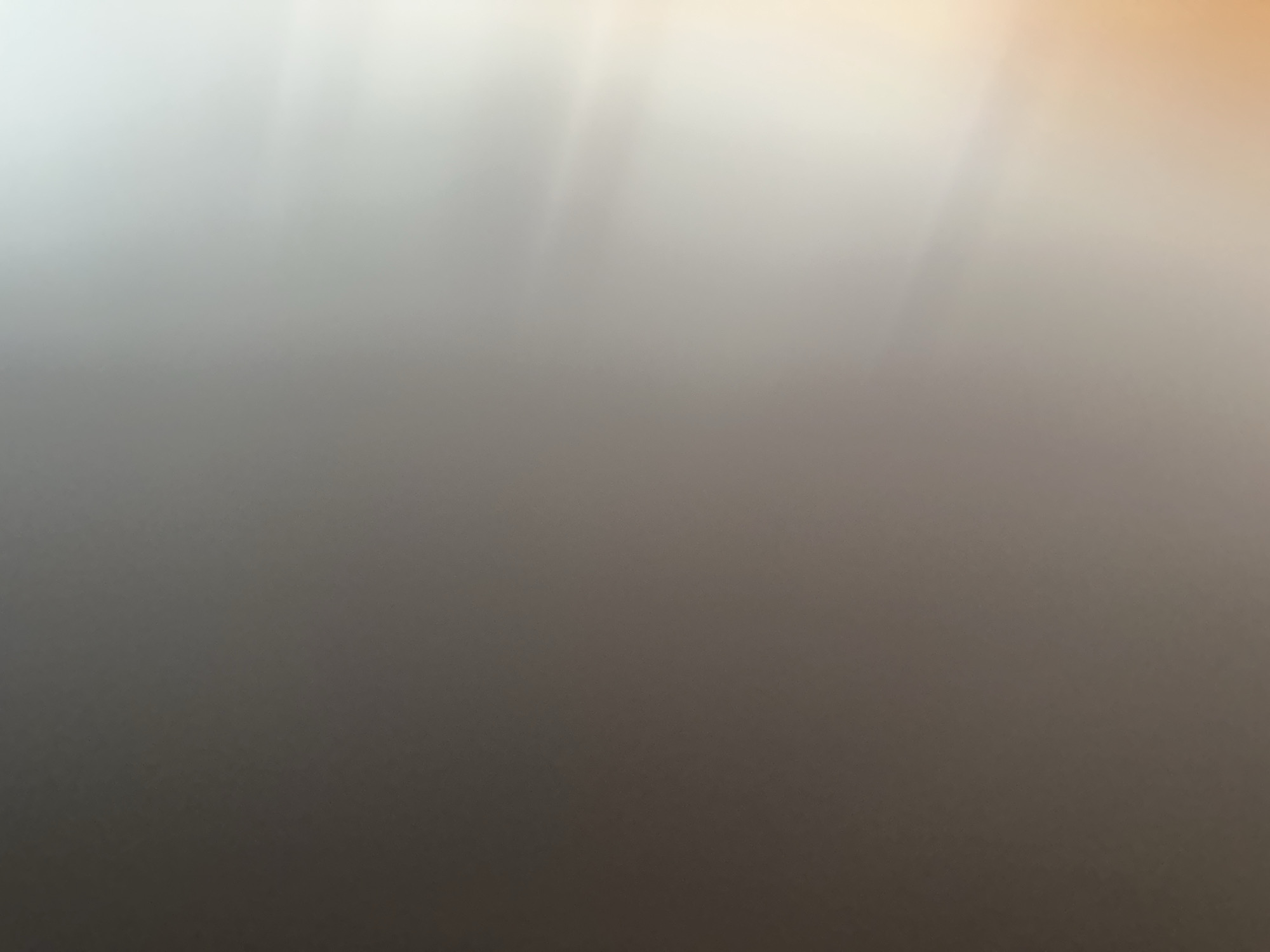 This is an abstract, out-of-focus image with a gradient of light and dark tones. The top of the image features soft, diffused light with a slight warm, orange hue, transitioning into lighter shades of gray and white. Vertical streaks of light are visible, suggesting a light source from above and to the right. The bottom portion of the image is a dark, muted gray, with the color becoming progressively deeper towards the bottom edge.
There are no discernible subjects, objects, scenes, or activities. The image appears to be a capture of light and shadow, possibly from a textured surface or through a translucent material. There are no visible text elements. The abstract nature of the image prevents specific details about time of day, weather, location cues, or emotions from being inferred. Given the provided location context of Belgrade, Serbia, it's possible this image is a stylistic representation of light within an urban or indoor environment, but without more information, this remains speculative. The overall impression is one of subtle color gradation and diffused light.