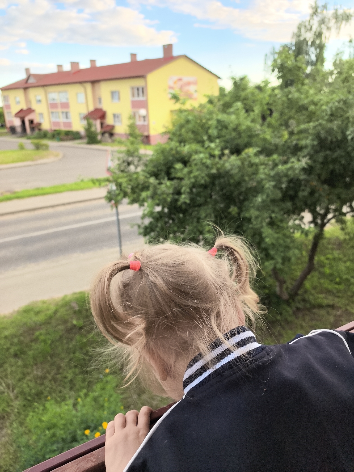 A person with blonde hair styled in two pigtails, secured with red hair ties, is seen from behind, leaning over a railing. They are wearing a black jacket with white and blue striped trim on the collar. In the background, a yellow apartment building with a red roof is visible, alongside a road and lush green trees. The sky is blue with scattered clouds.